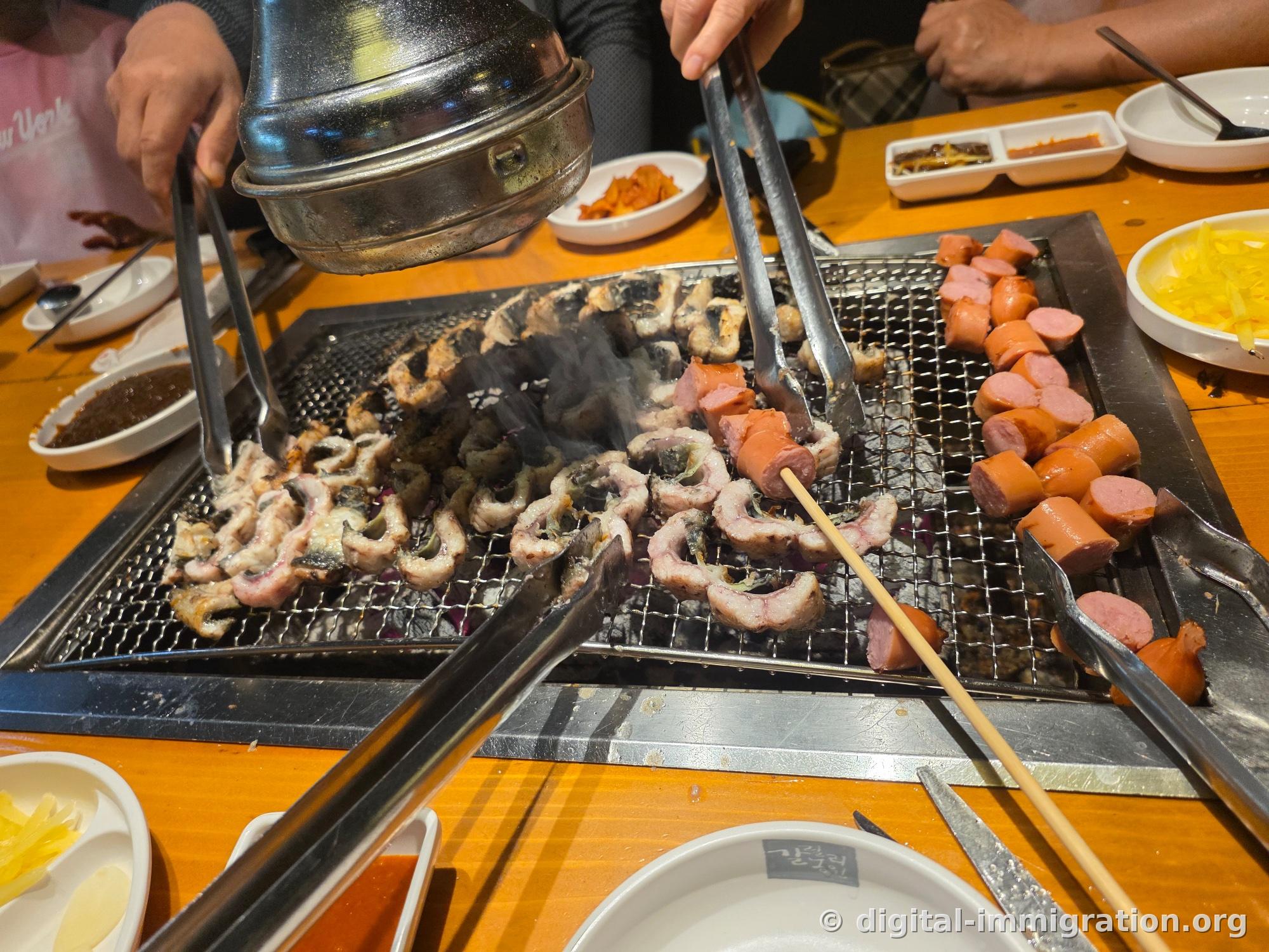 La grille pleine de morceaux d'anguille coupés à la taille d'une bouchée qui se recourbent et cuisent, avec des saucisses tranchées qui cuisent à côté. Deux pinces retournent les anguilles