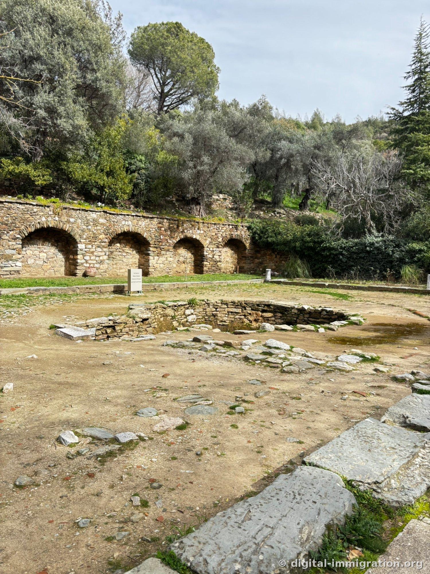 An excavated pool-shaped stone structure is revealed in front of a low stone wall with four arches carved through, with silvery olive trees covering the hillside behind