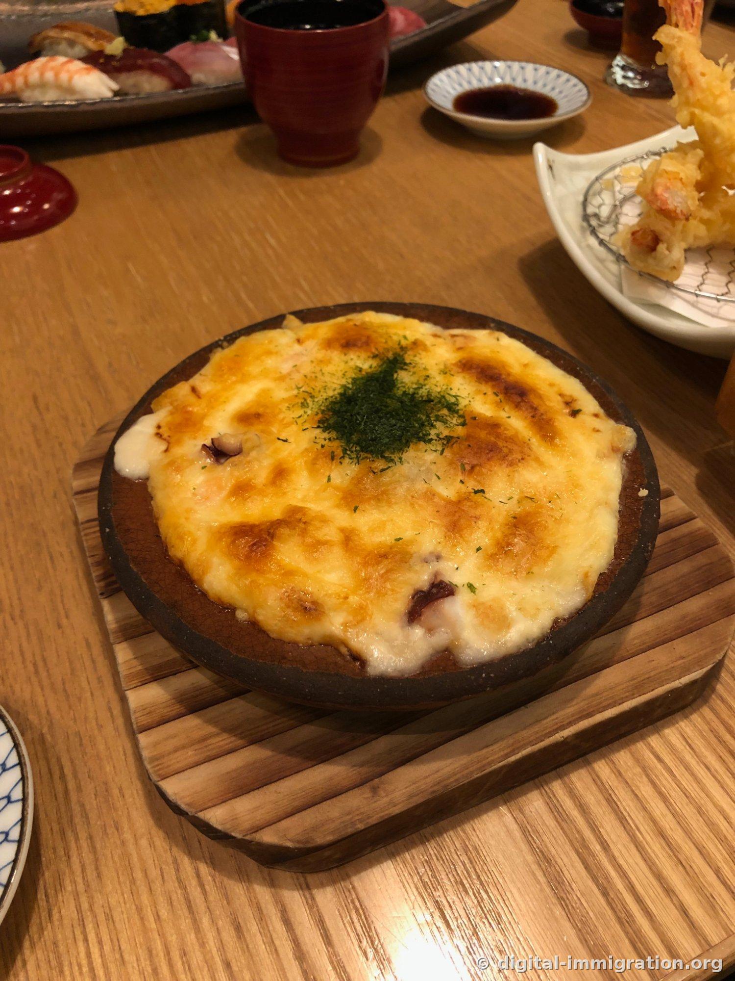 Gratin de fruits de mer dans un bol en céramique sur un support en bois. Le fromage est bruni et de la poudre d'algue nori est saupoudrée au milieu