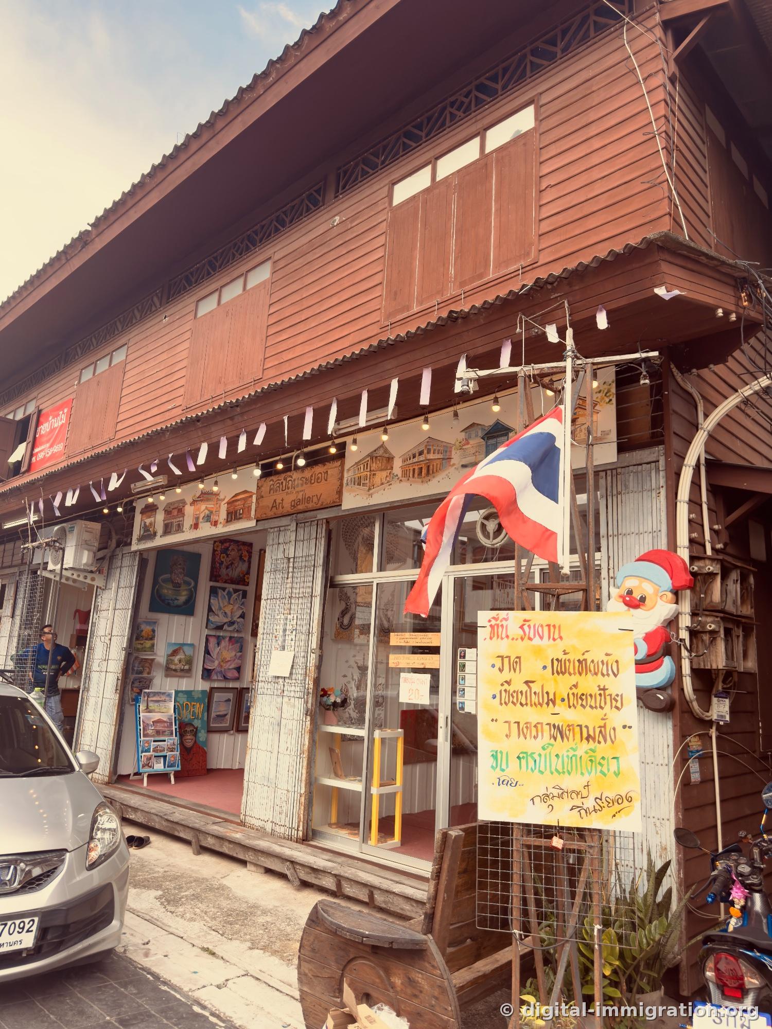 Art gallery storefront on ground floor of wooden two-story building with displayed paintings and Thai flag, Santa Claus decoration