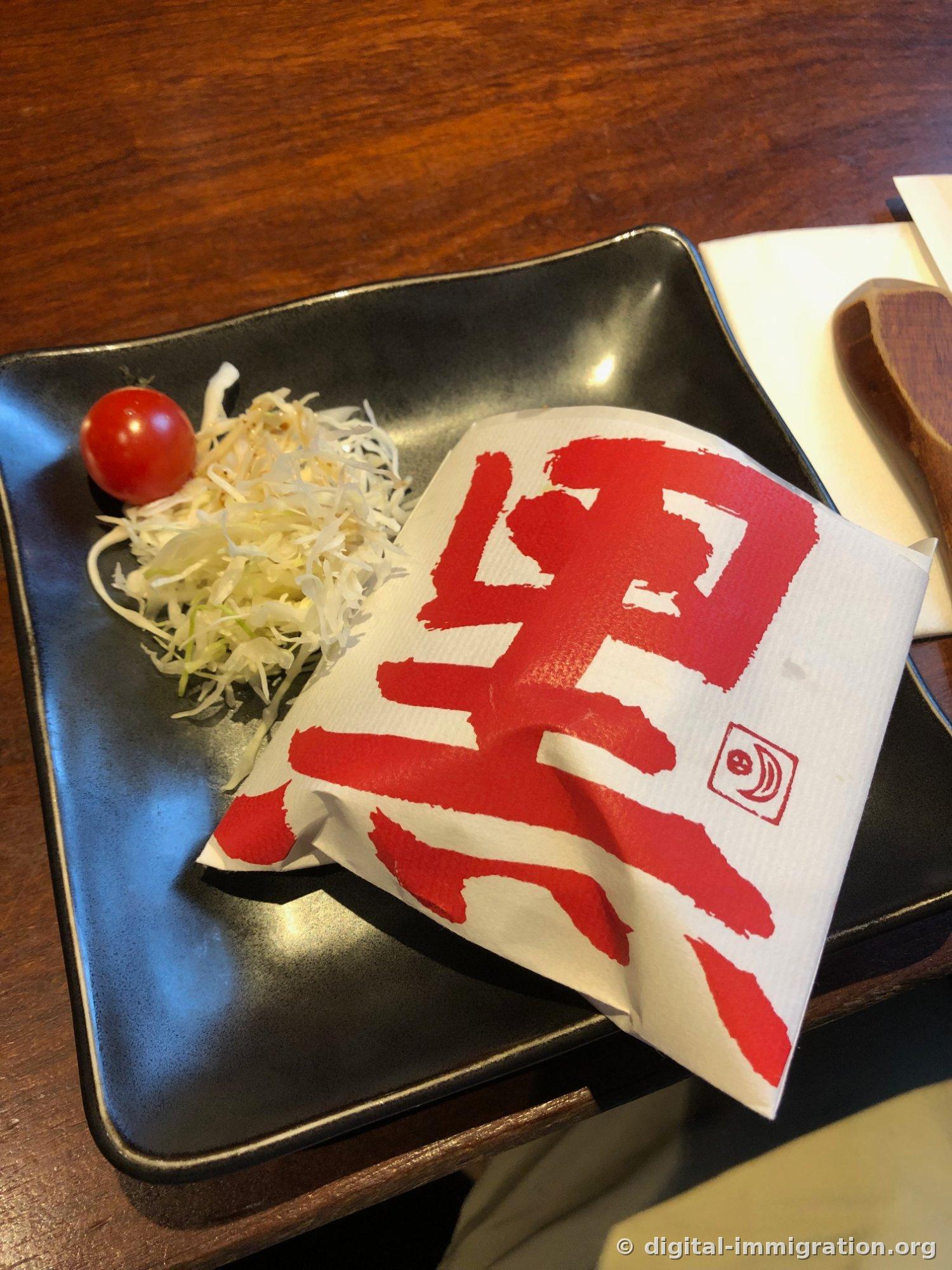 黒い角皿にキャベツの千切りとミニトマトが置かれ、赤い筆書き「黒」の字が大きく印刷された紙が一緒に敷かれている様子