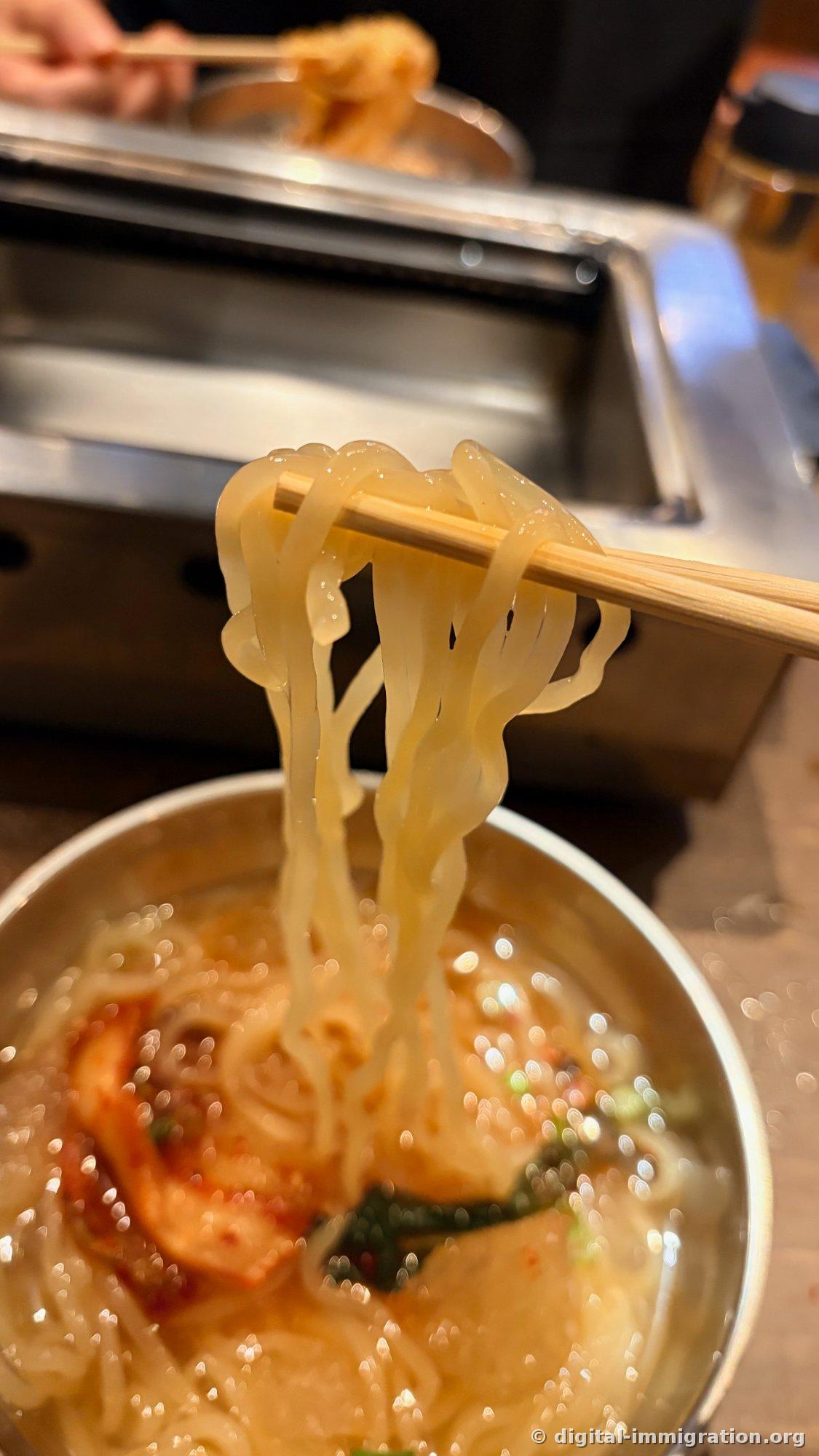 Thick, translucent noodles lifted with chopsticks; the bowl below holds kimchi and broth, with the stainless-steel roaster visible in the background