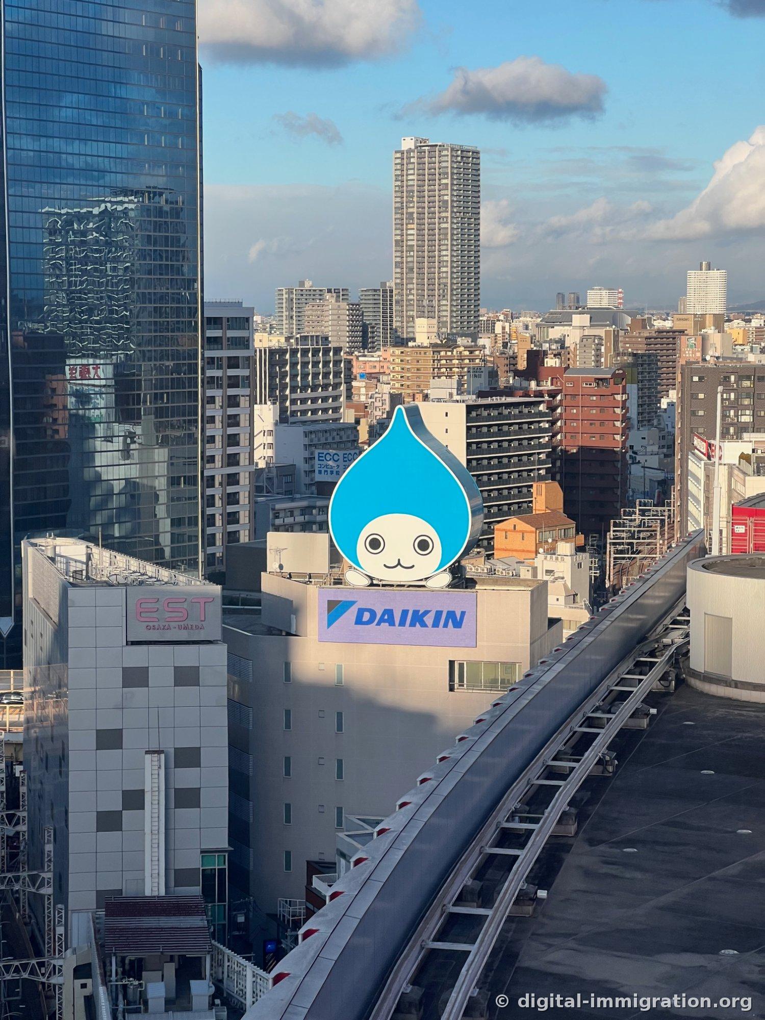 観覧車レールの向こうに、ダイキン屋上看板と青い水滴キャラクター、左側にEST OSAKA-UMEDA看板が見える梅田市街地