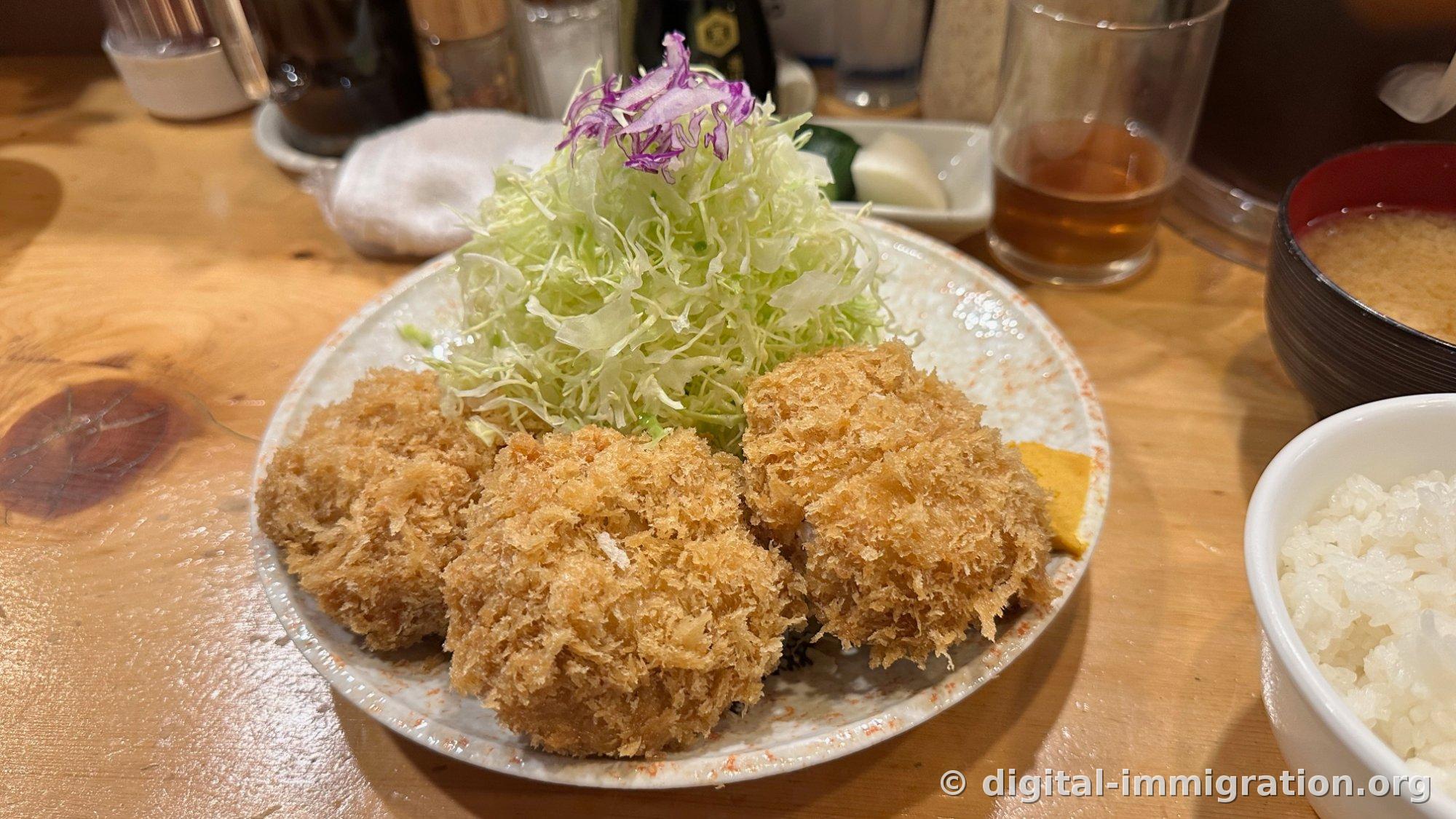 Three filet cutlets on a white plate with shredded cabbage piled high like a mountain, rice and miso soup on a wooden table