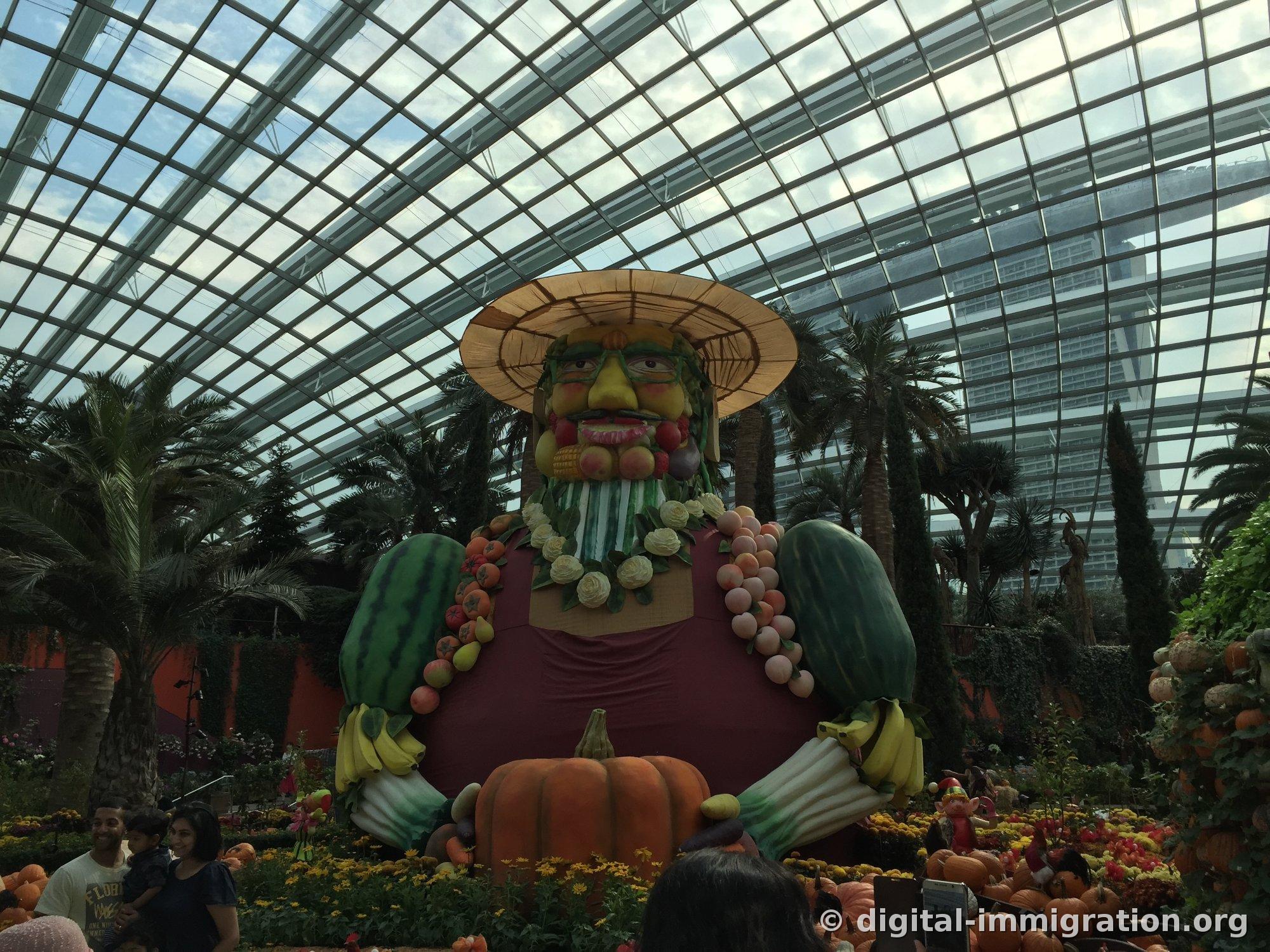 The giant figure wearing a straw hat viewed from the front, with a face made of fruit and a beard made of vegetables, with the Marina Bay Sands building visible beyond the glass roof