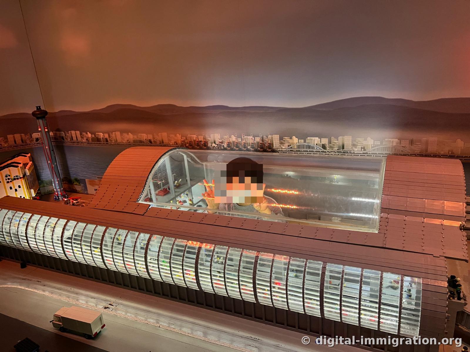 アーチ型の透明な屋根で覆われた大型駅舎のレゴ模型で、屋根の隙間から子どもが中を覗き込んで手を振っている