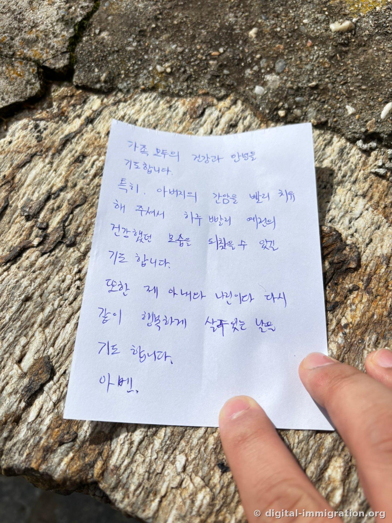A white sheet on rough stone with a Korean prayer written in blue ballpoint pen, contents asking for family health and father's healing, with two fingers holding the paper down