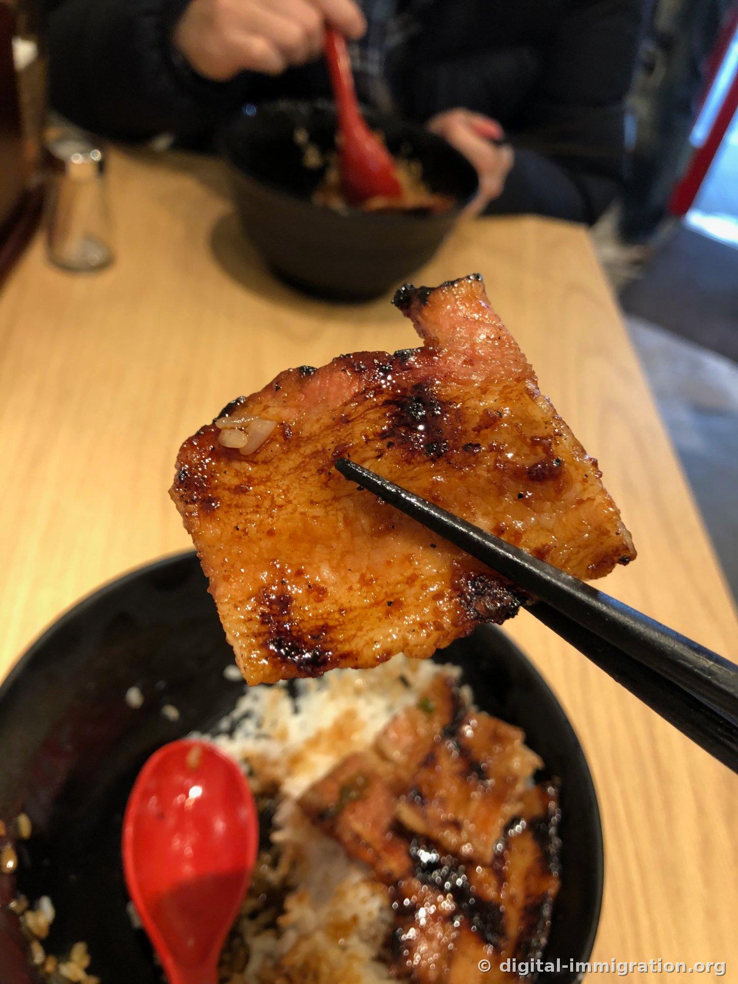 Un morceau épais de porc grillé soulevé par des baguettes noires. En dessous dans le bol, du riz et de la viande restante, ainsi qu'une cuillère rouge en plastique, et un autre bol identique de l'autre côté