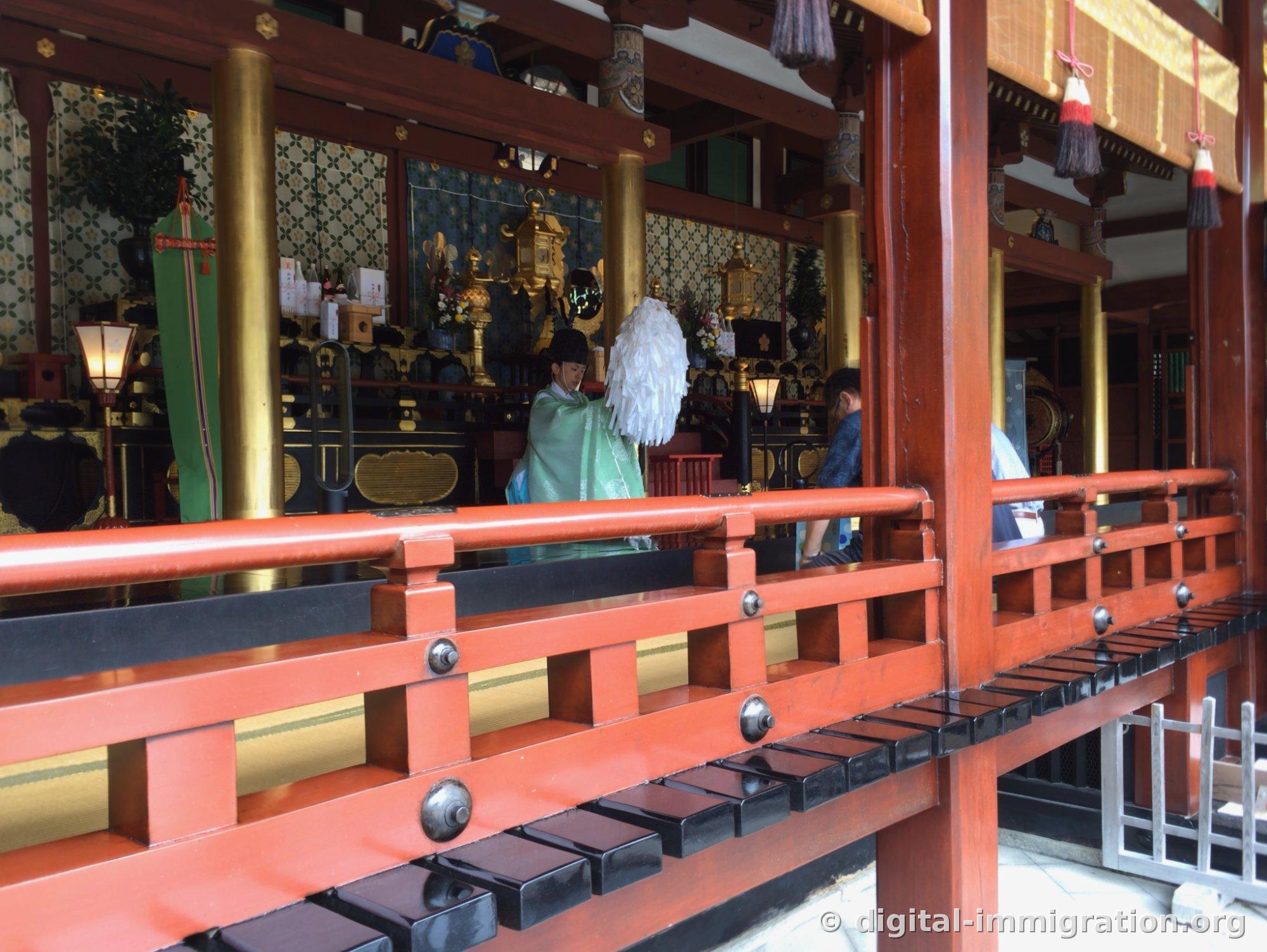 Priest in pale green robes holding an onusa with white paper inside the main hall, conducting a ritual
