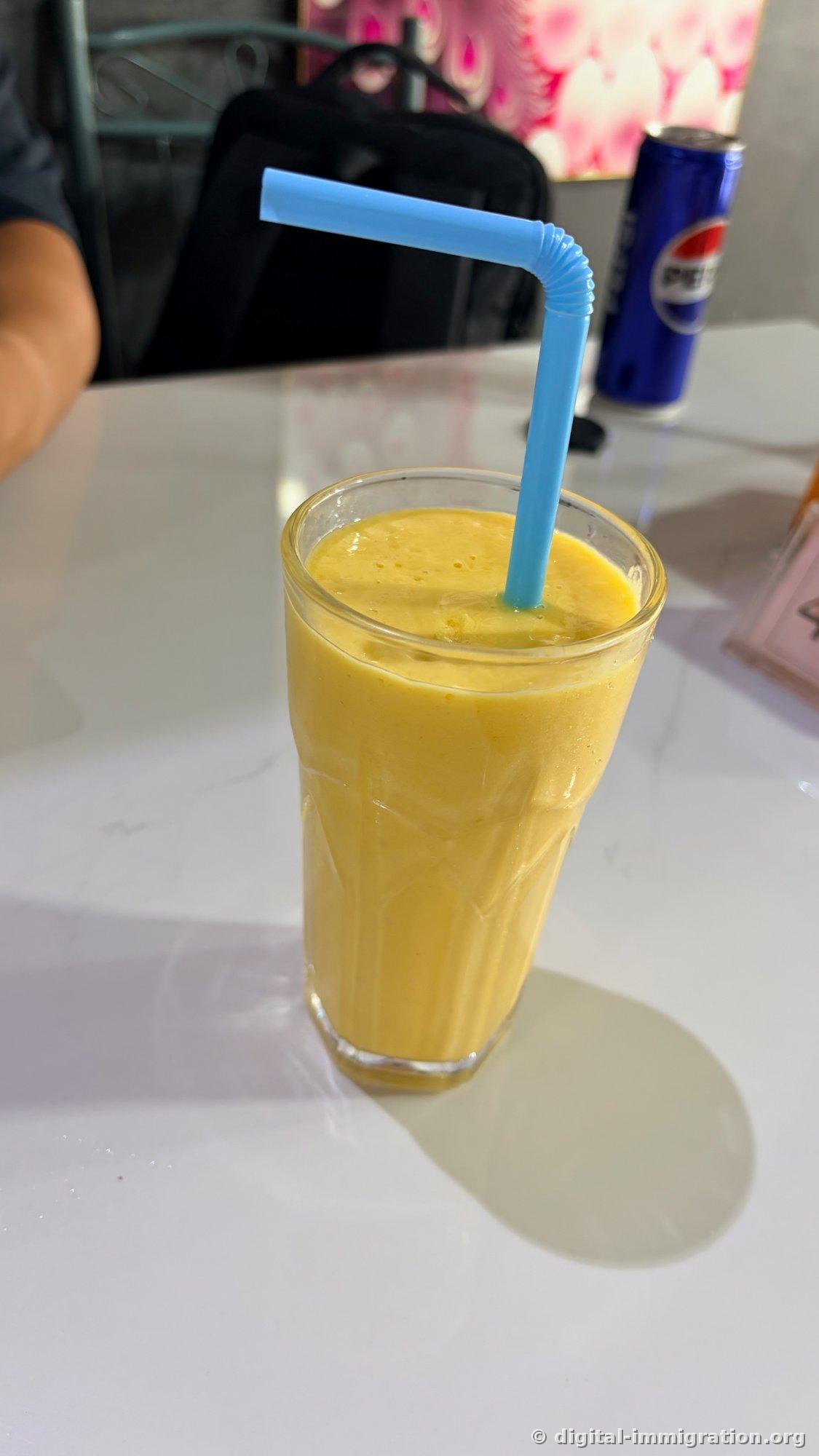 Lassi à la mangue jaune foncé dans un grand verre avec une paille bleue, avec une canette de Pepsi bleue visible à l'arrière-plan sur la table