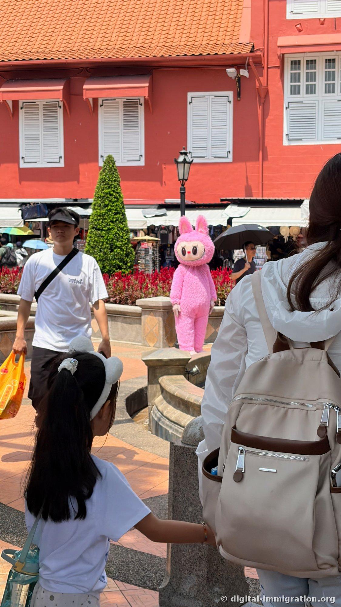 A person in a pink rabbit mascot costume stands in a square in front of a red building, and a young girl looks toward them while holding an adult's hand. People with umbrellas are visible in the shop area behind
