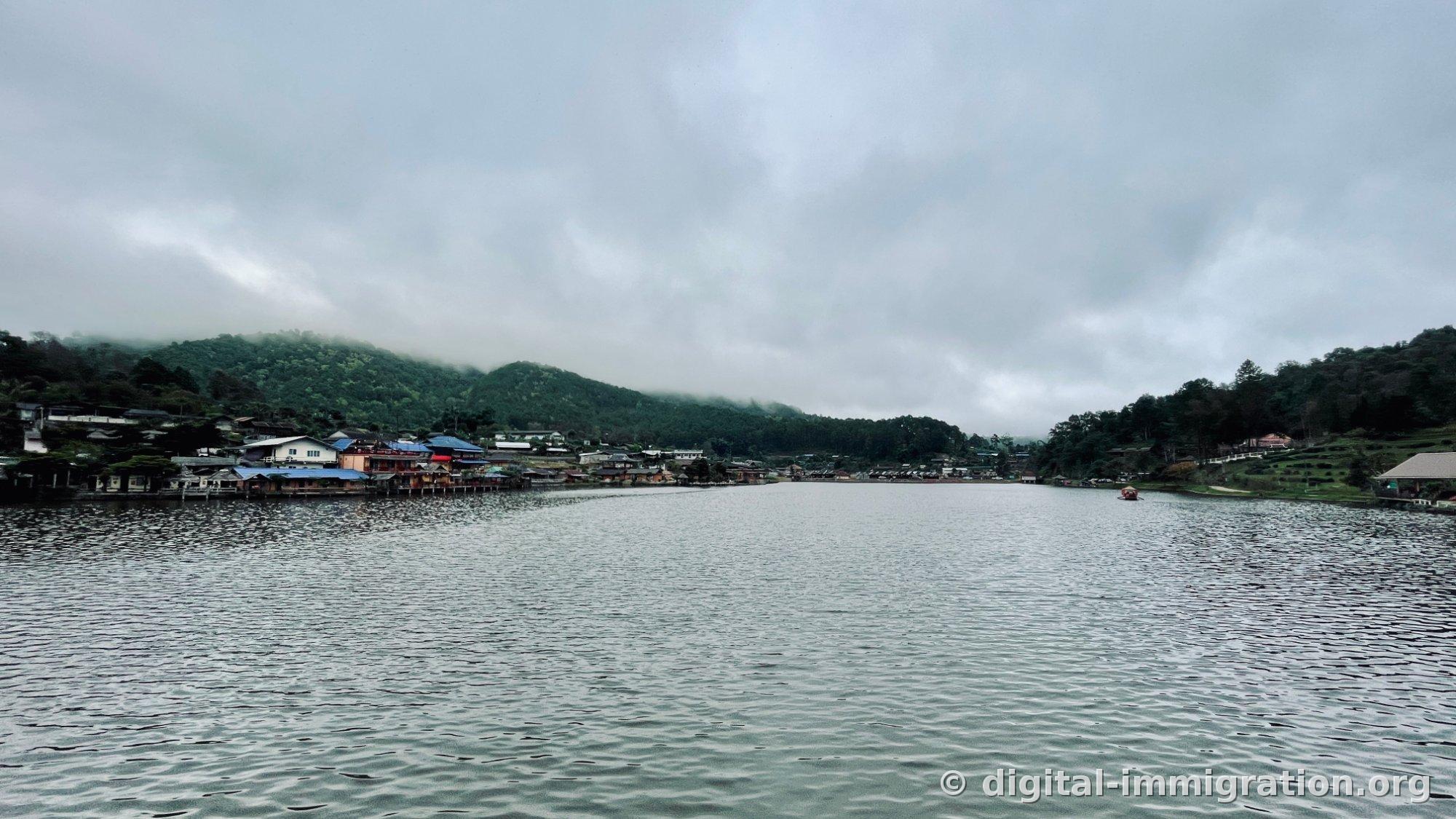 Le lac Ban Rak Thai par temps nuageux, avec des nuages gris s'accrochant à la crête de la montagne. Des petits cafés aux toits bleus s'alignent le long de la rive gauche