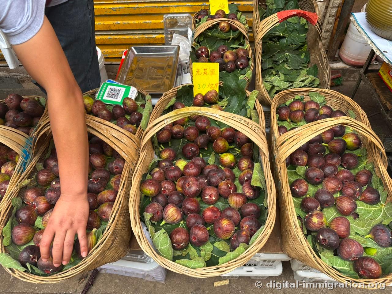 Dark purple figs heaped neatly in a bamboo basket lined with fig leaves, yellow price sign reading '无花果 20元/公斤', vendor's hands arranging the basket