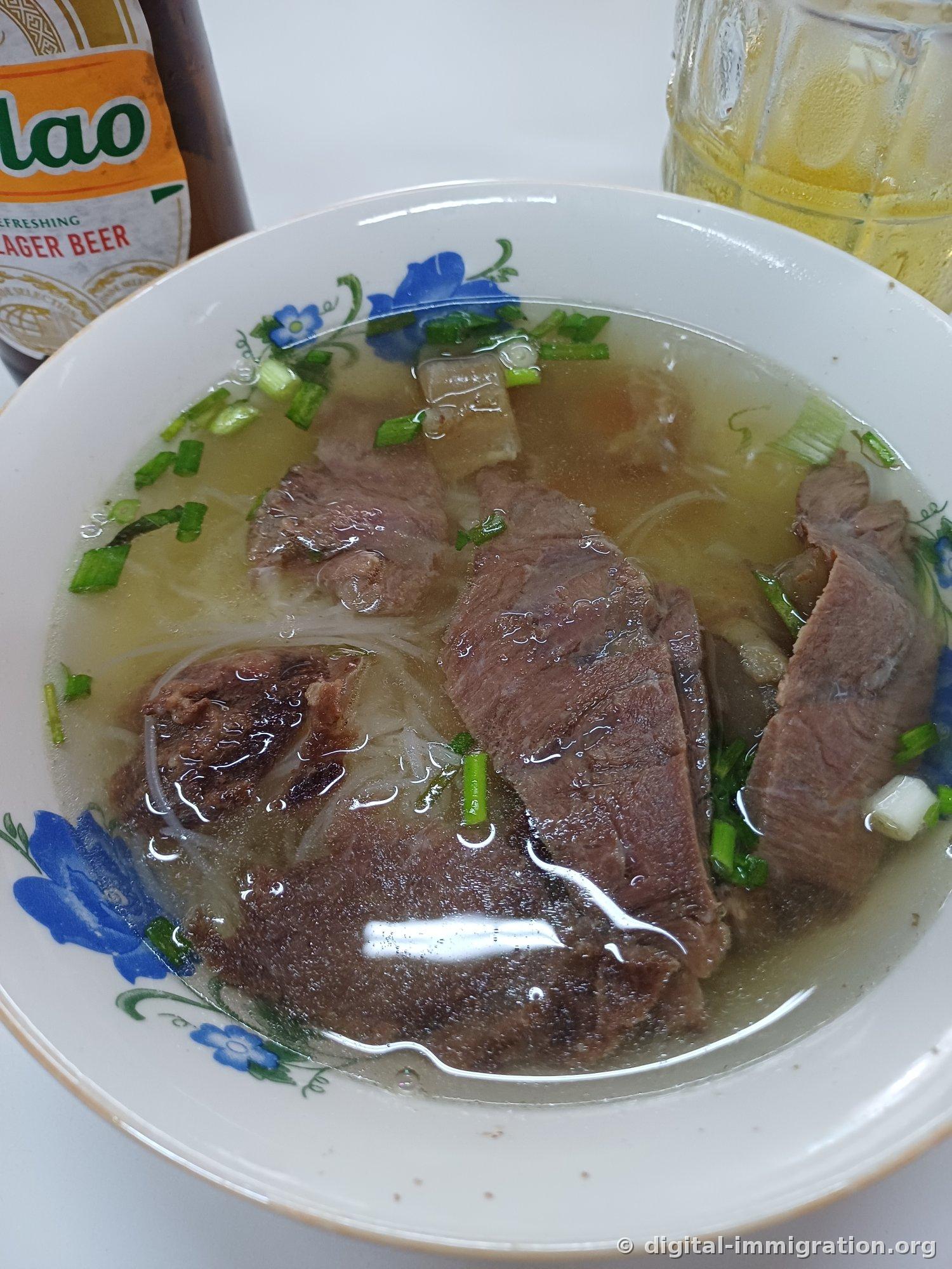 花纹陶瓷碗里盛着清汤牛肉米线，有块状牛肉和透明的牛膝软骨，撒上葱段，背景是啤酒瓶和冰水杯