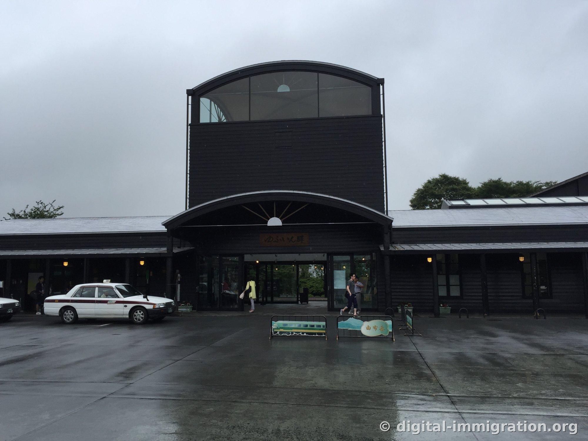 Au-delà du rond-point mouillé par la pluie, la façade de la gare de Yufuin en bois noirci, avec un taxi blanc en attente