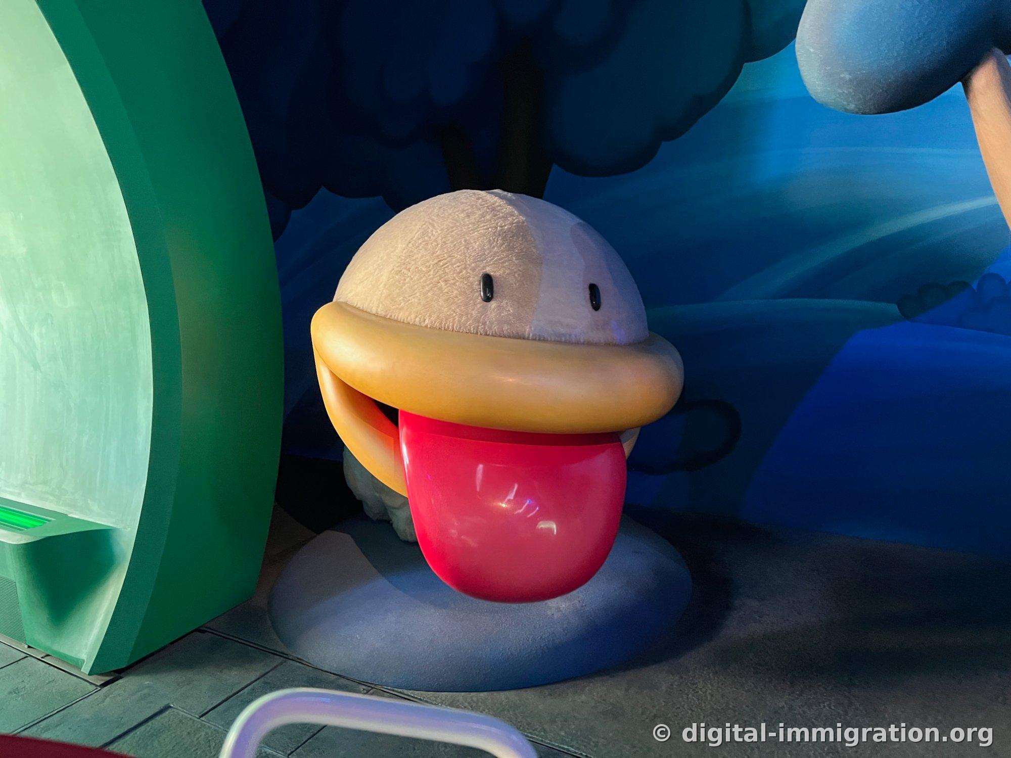 A character sculpture on the floor of a dark indoor section with white fur-textured head, yellow lips, and a large extended red tongue
