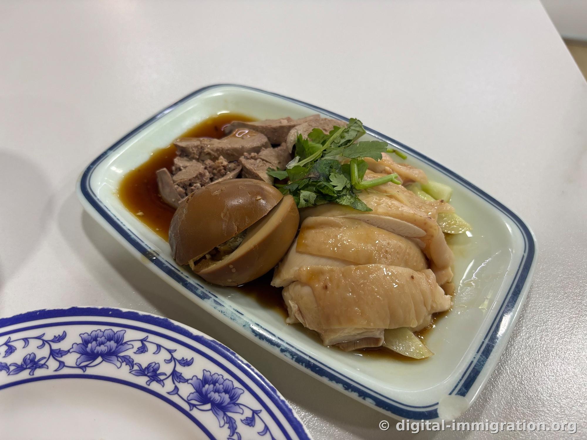 Blue and white porcelain plate with sliced poached chicken, half a soy-braised egg, chicken liver pieces, and cilantro. Thin soy sauce pools at the bottom