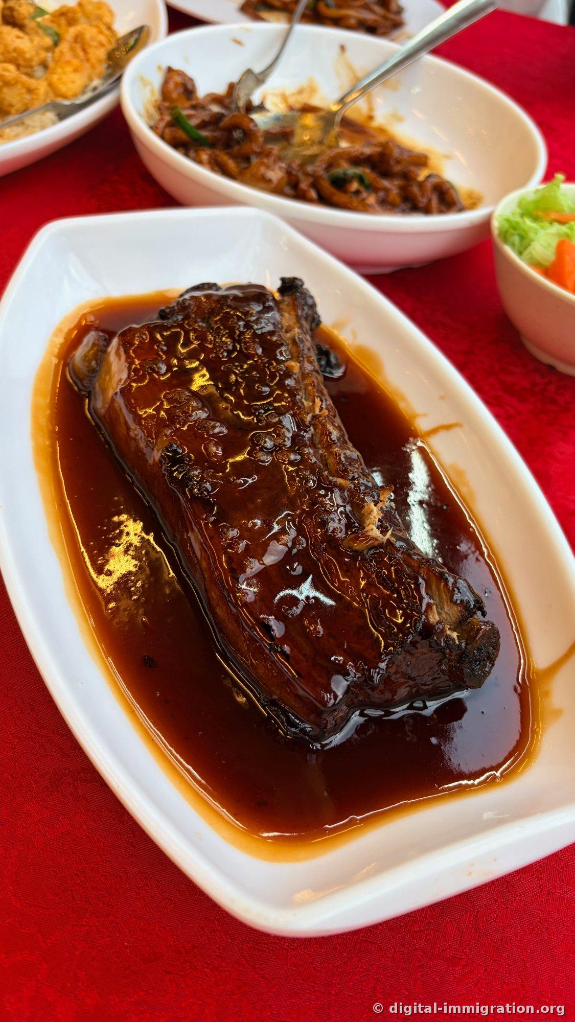 深紅色のソースに浸った豚バラ肉または短いあばら骨の塊が白い楕円形のお皿に置かれ、後ろにホッキエン麺のボウルと野菜の漬物、カレー揚げのお皿が一緒に置かれたテーブル
