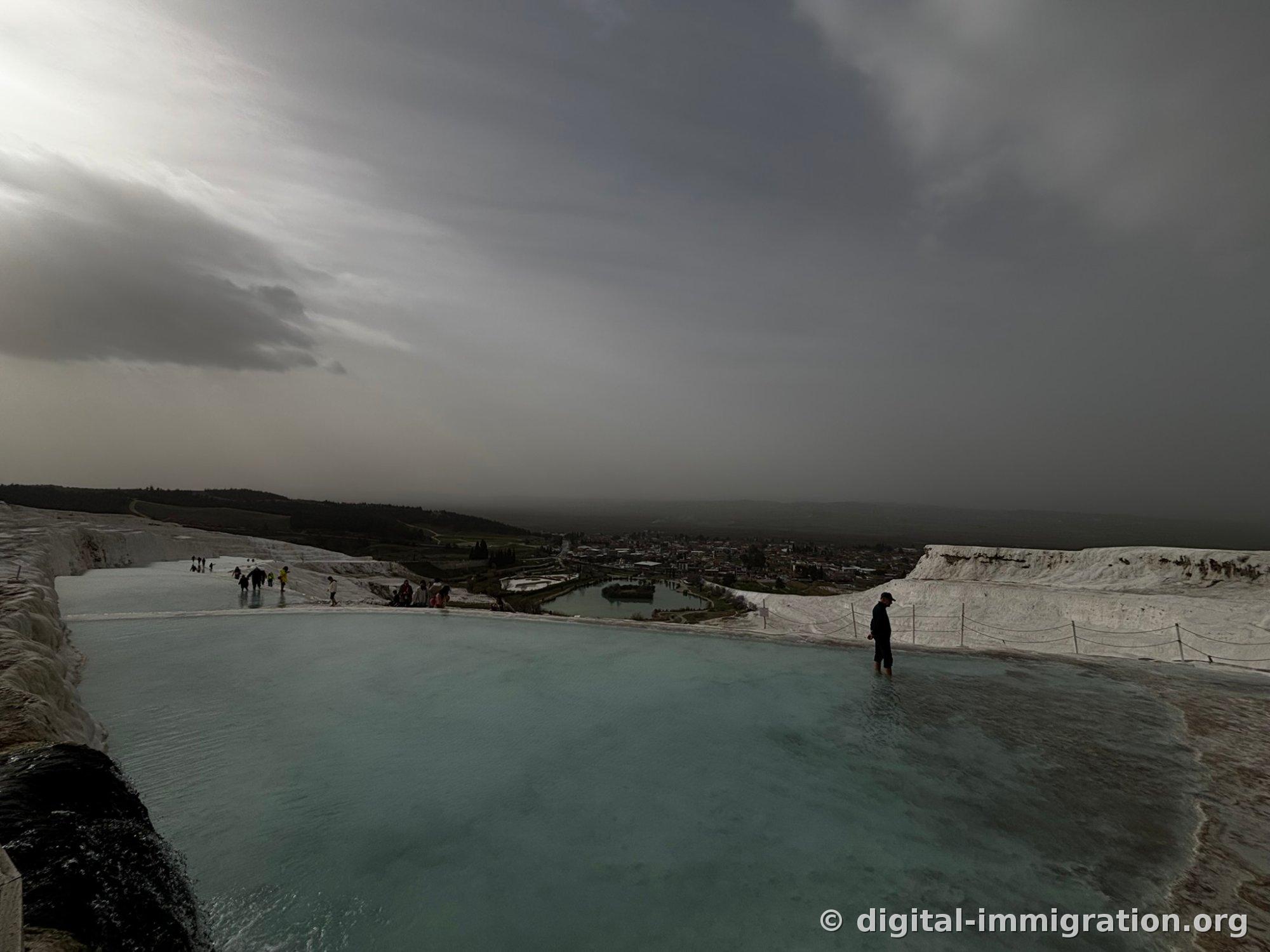 An eerie landscape—one person standing ankle-deep in a wide limestone travertine puddle beneath heavy dark clouds and a low gray sky