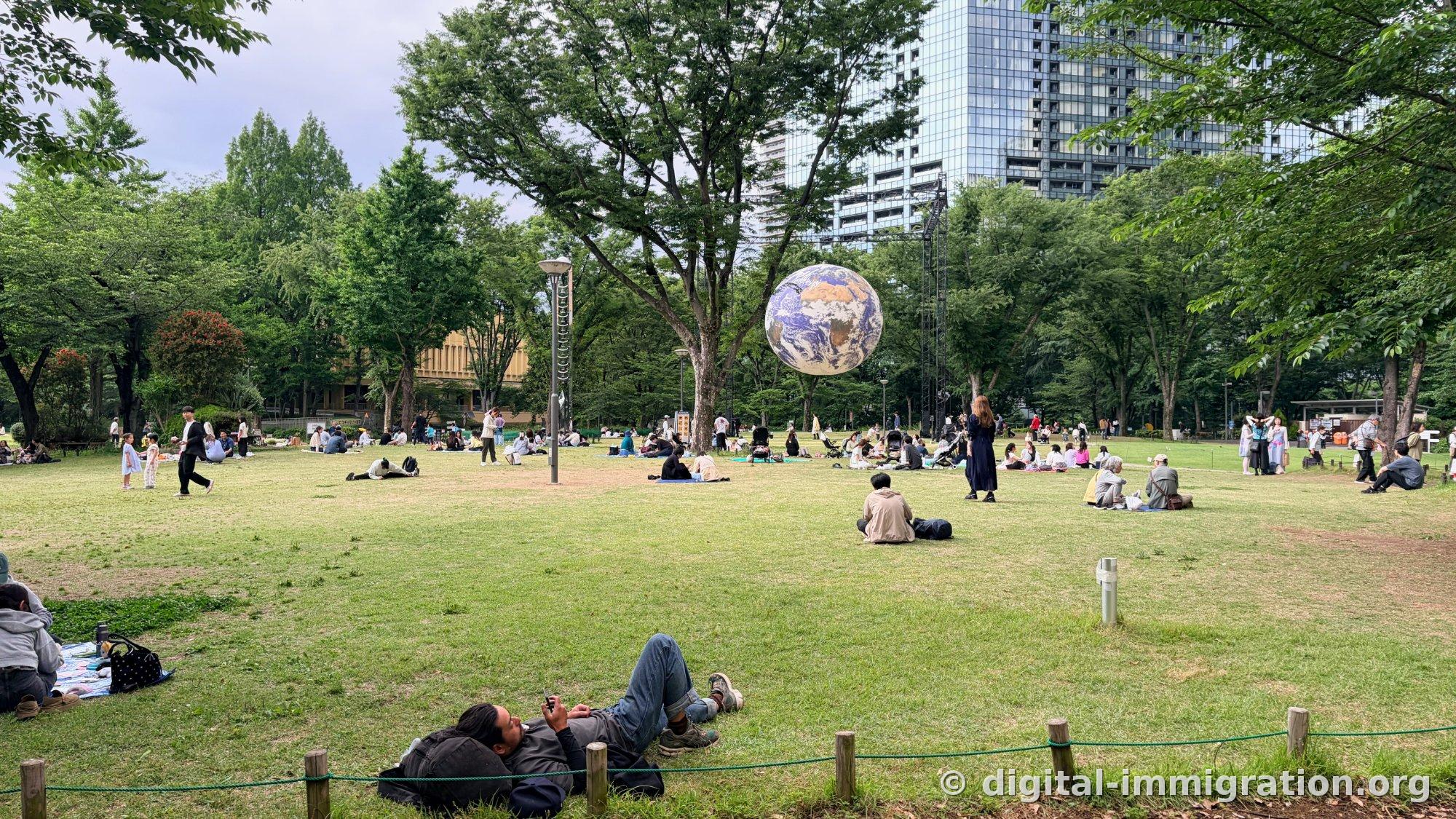 Sculpture de globe suspendue dans la pelouse du parc central de Shinjuku avec des gens assis sur des nattes ou allongés en dessous, avec des gratte-ciel en verre à l'arrière