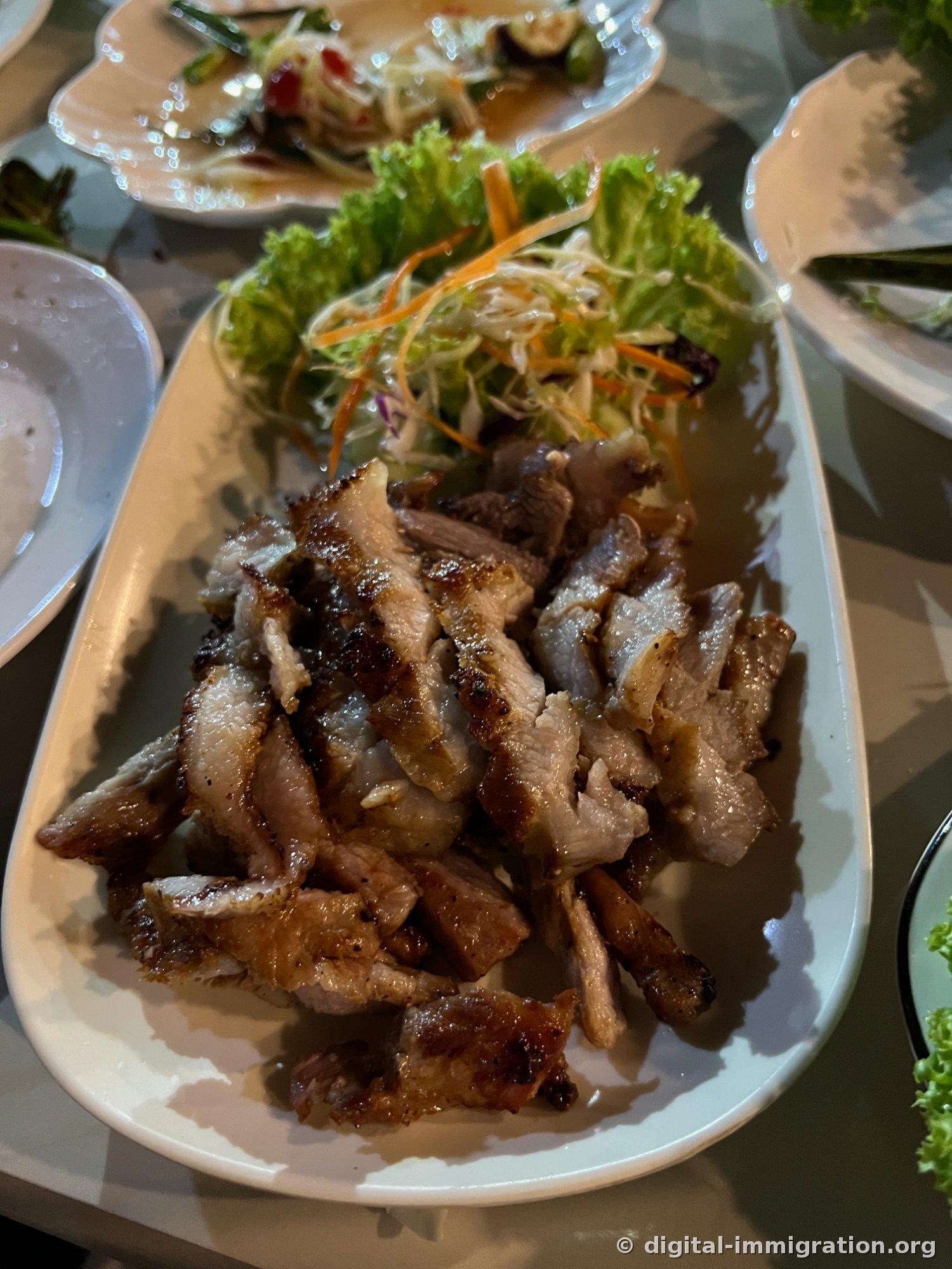炭火烤至斜切的猪肉盛在白色椭圆盘上，上面铺着切丝的卷心菜、胡萝卜和生菜
