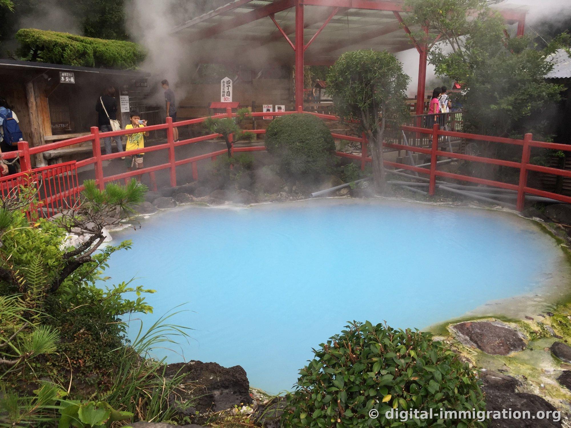 Un bassin d'eau chaude bleu ciel entouré d'une rambarde rouge et une pancarte lisant '四丁目 かまど地獄 80℃', avec de la vapeur s'échappant