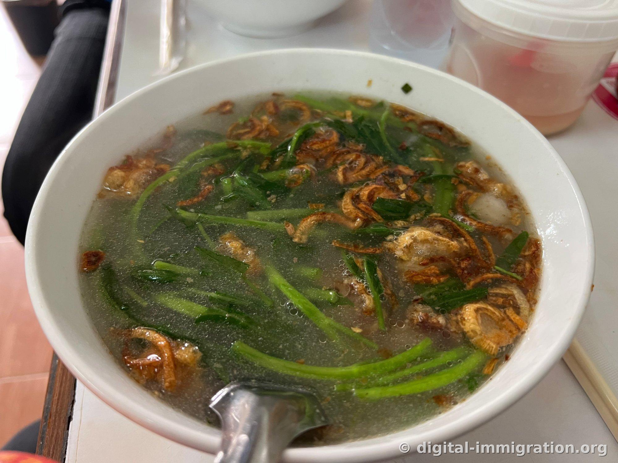 Khao poon noodles in a white porcelain bowl, topped with fried garlic and green vegetable stems over thick broth, with fine foam floating on the surface