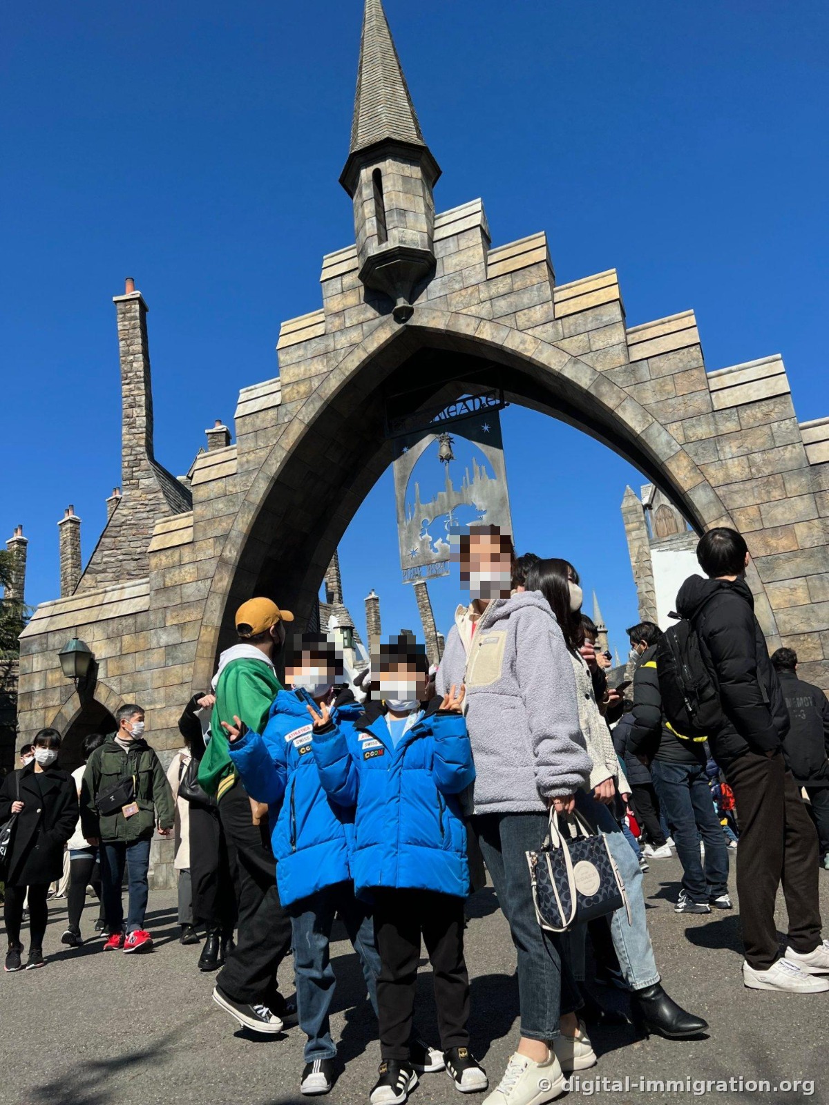 Children in blue padded jackets make peace signs beneath the pointed stone arch at the Hogsmeade entrance, surrounded by crowd-wearing visitors