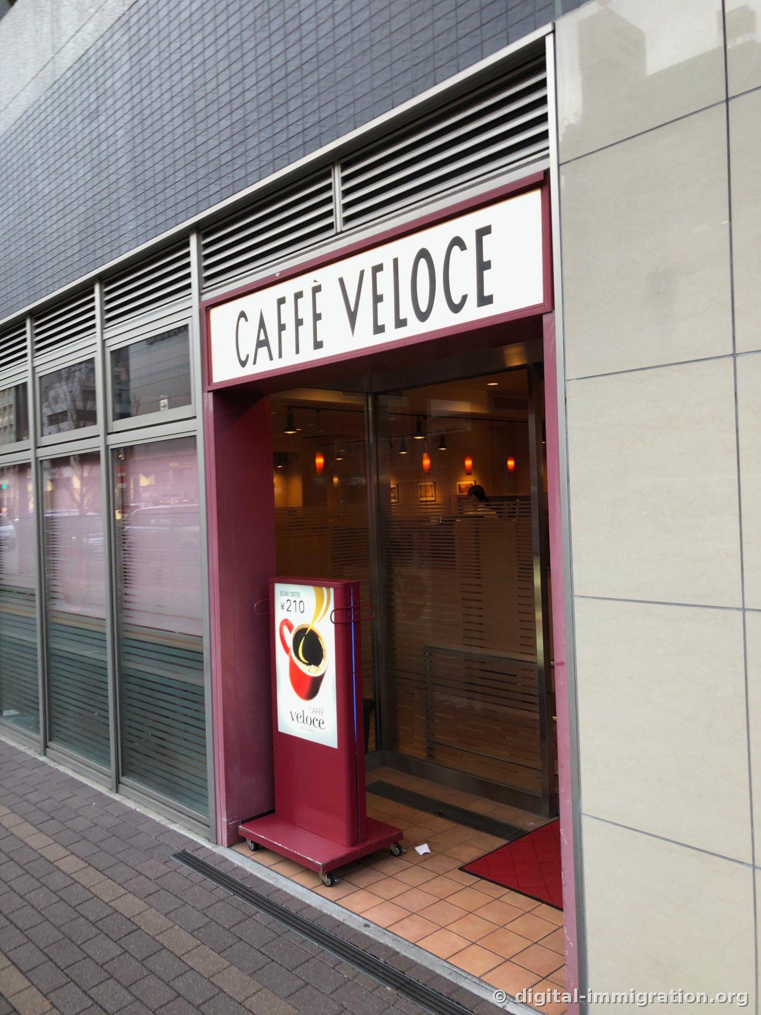 L'entrée du CAFFÈ VELOCE avec un cadre rouge vin au premier étage d'un immeuble en carrelage gris, et une pancarte à roulettes indiquant 'café mélange ¥210'