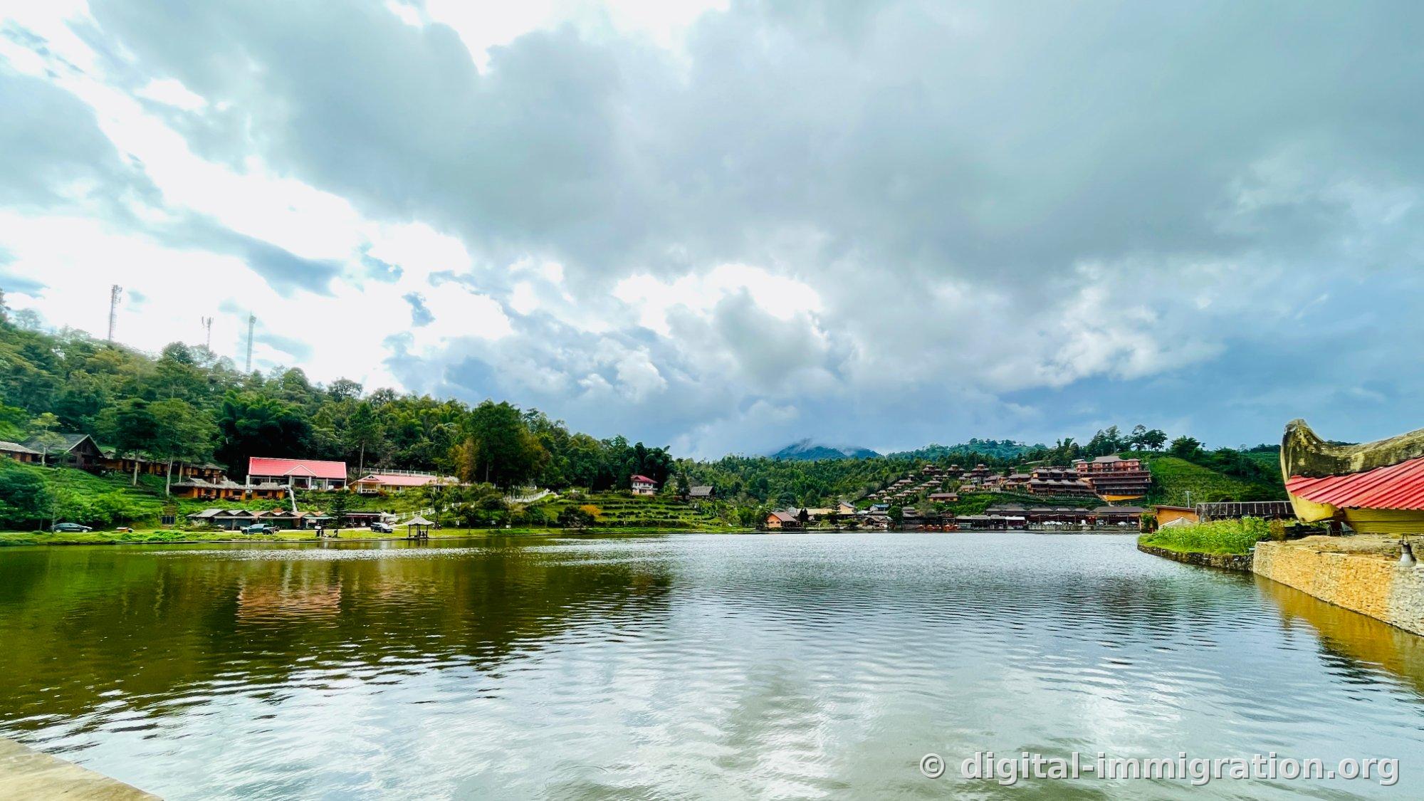 Vue d'ensemble du village Ban Rak Thai depuis l'autre côté du lac. Un bâtiment aux toits rouges sur la colline gauche, un complexe de villégiature en bois sur la colline droite, et une structure en forme de bateau à l'avant droit