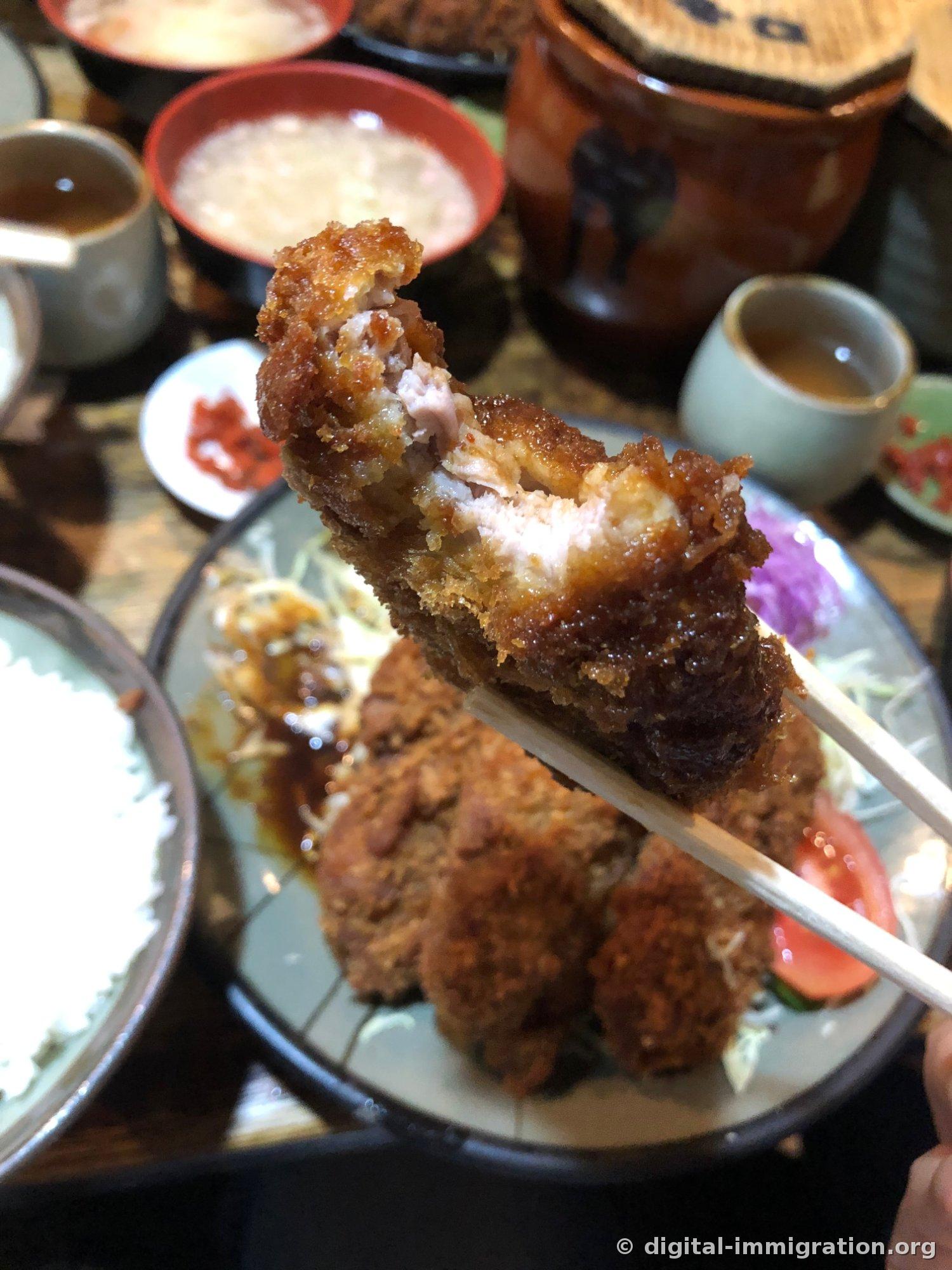 Cross-section of a tonkatsu piece held up with chopsticks, showing white meat grain visible inside the dark brown batter