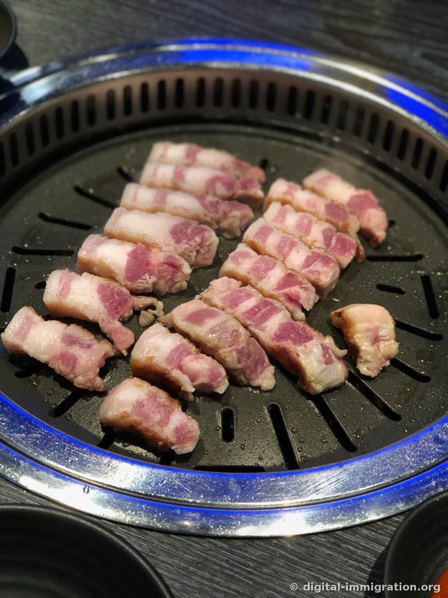 青いリングで囲まれた円形の焼き板の上で焼かれている厚切りのサムギョプサルの切れ端が、端が褐色に焼け始めている様子