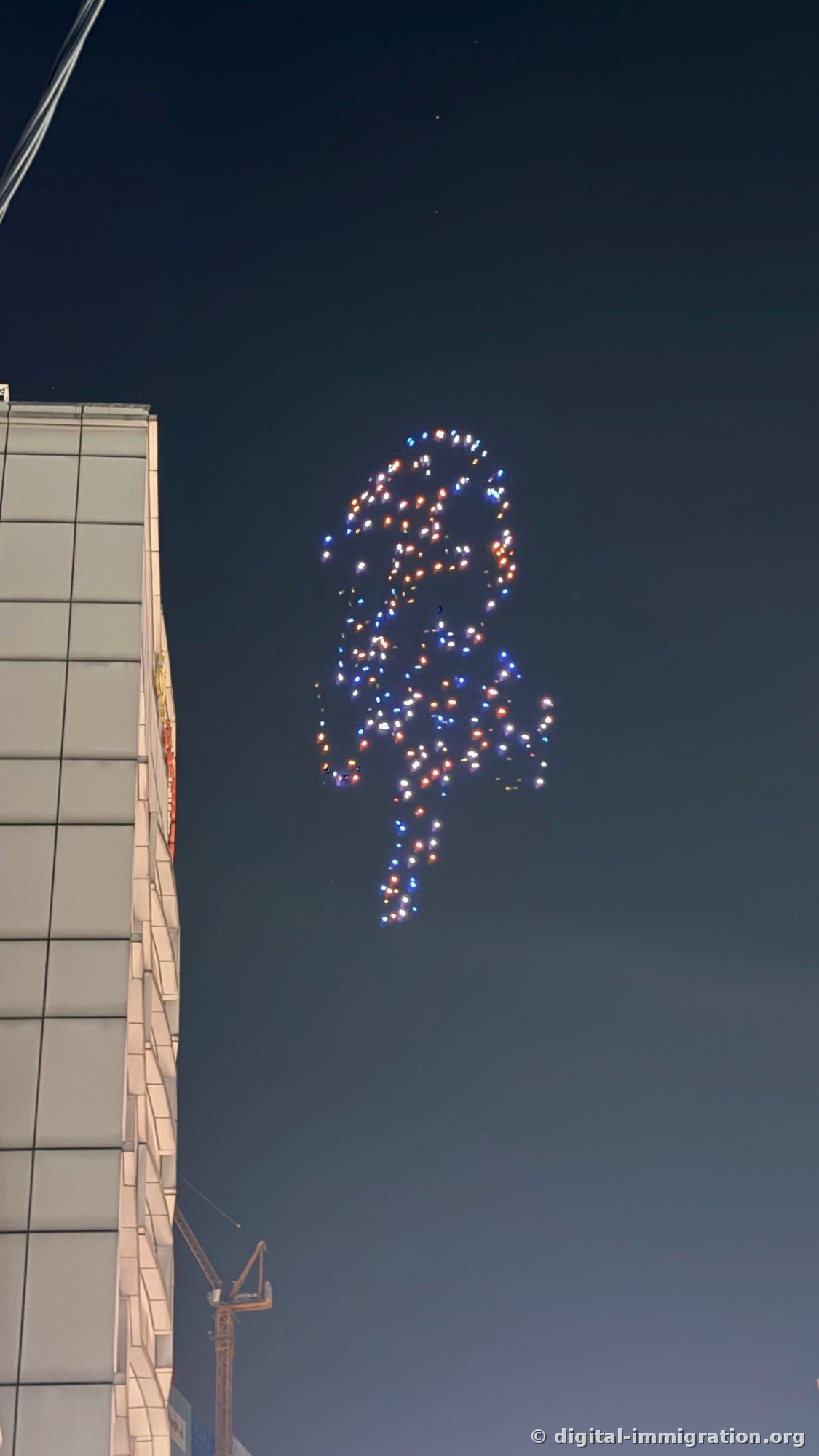 暗くなった空に赤と青の照明を点けた数百台のドローンが集まって形を作っており、左側に高層建物の外壁と下の方にタワークレーンが見える光景