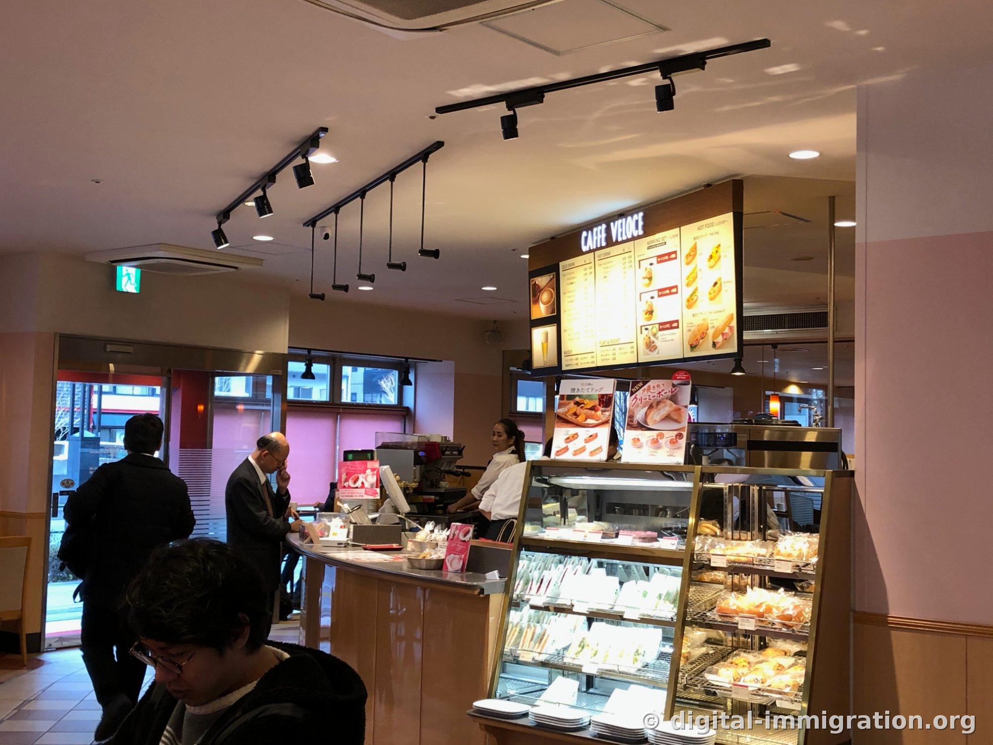Vue d'ensemble du comptoir de commande à l'intérieur du magasin. Le plafond est équipé d'éclairage sur rail noir en succession, sous le panneau de menu jaune avec le logo CAFFE VELOCE, il y a une vitrine en verre remplie de sandwichs et de pains, et un client en costume attend la commande au comptoir
