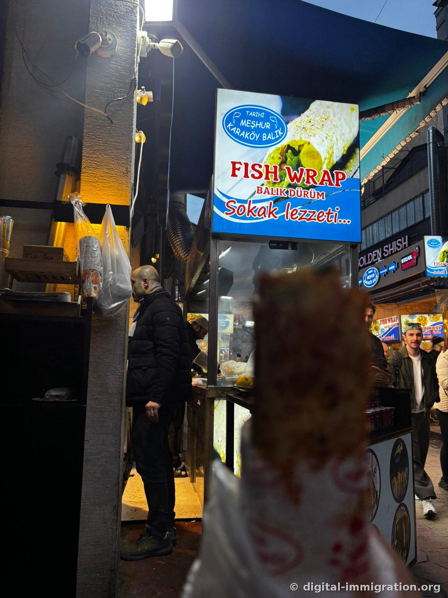 A mackerel wrap held in hand with the blue FISH WRAP BALIK DÜRÜM sign and night street stall visible behind it