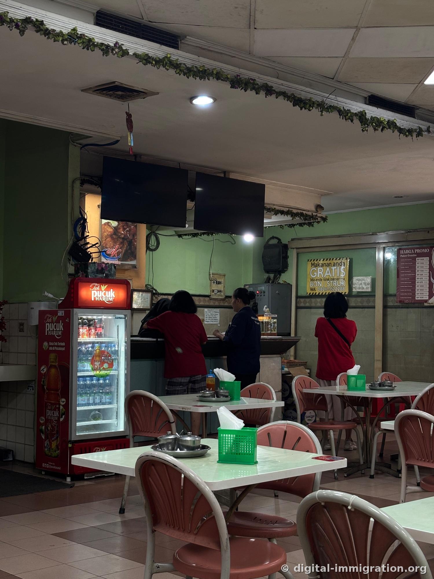 Intérieur du restaurant avec murs vert pâle et chaises en plastique rose, réfrigérateur de boissons Teh Pucuk Harum à gauche et employés derrière la caisse