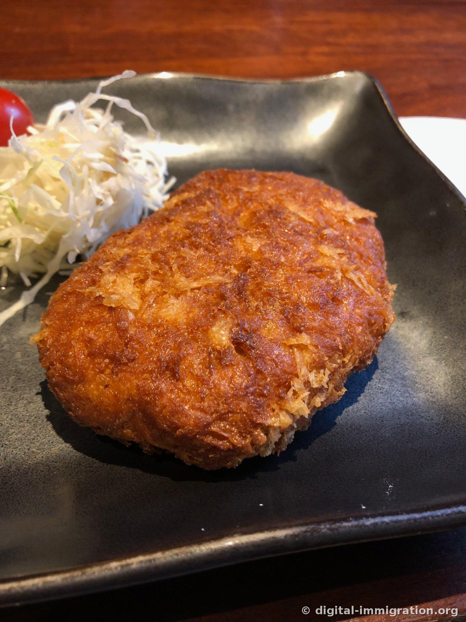 黒い角皿の上に濃い茶色に揚げられた楕円形のメンチカツ一枚とキャベツの千切り、ミニトマト