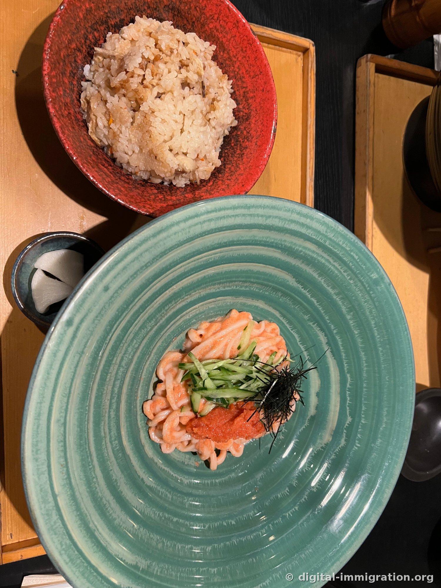 Udon à la crème de merlan sur un large plat bleu-vert. Les nouilles enrobées de sauce crème beige sont garnies de concombre tranché, d'œufs de merlan et d'algue nori finement râpée, avec un pot de riz cuit en cocotte dans un plat rouge et des radis marinés à l'arrière