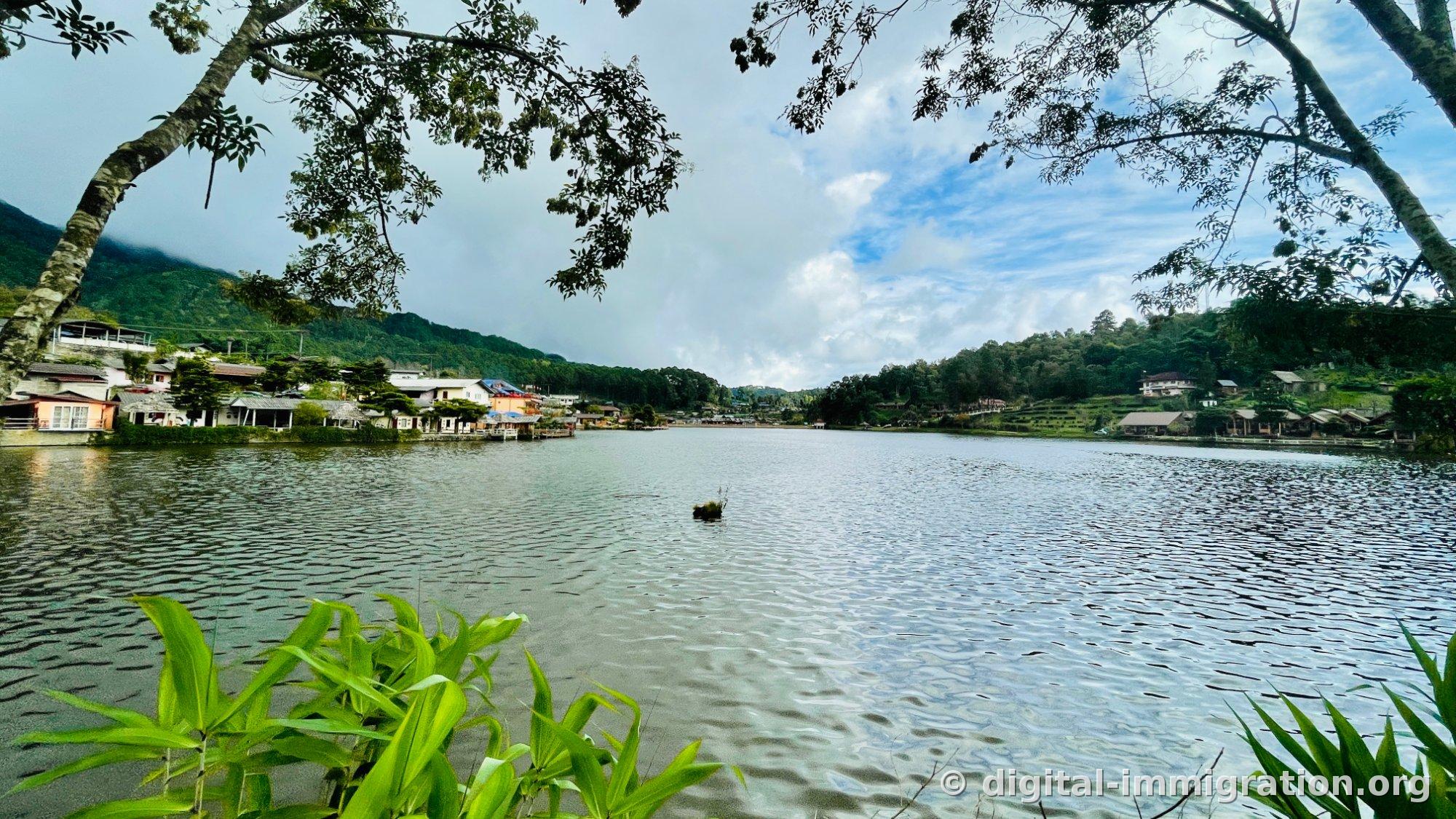 Le lac Ban Rak Thai par beau temps, vu à travers les branches des arbres. Des bâtiments orange et blancs s'alignent le long de la rive gauche