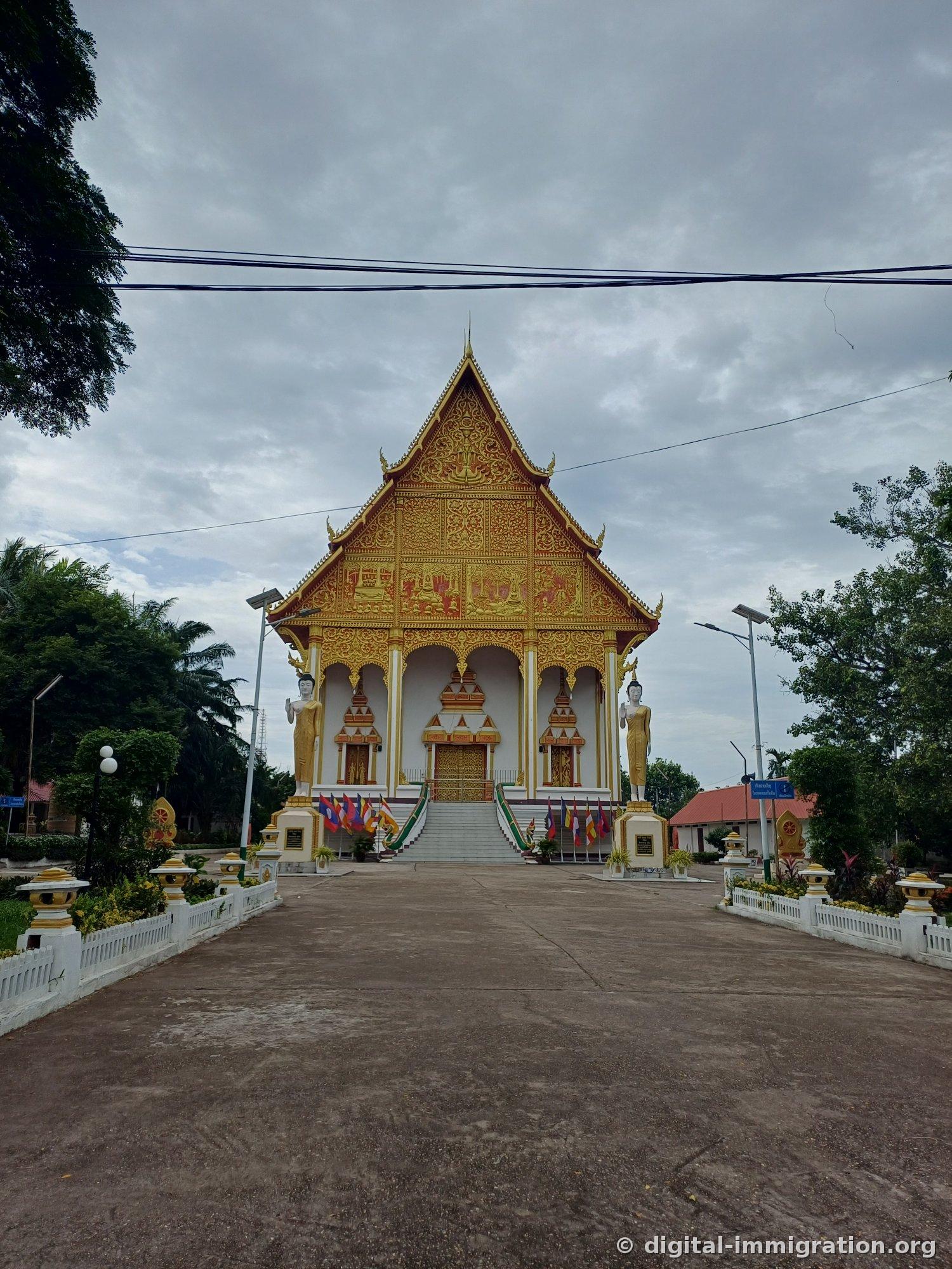 Front facade of ordination hall with Buddha life scenes carved into gilded gable, two Buddha statues on either side, Buddhist flags on staircase