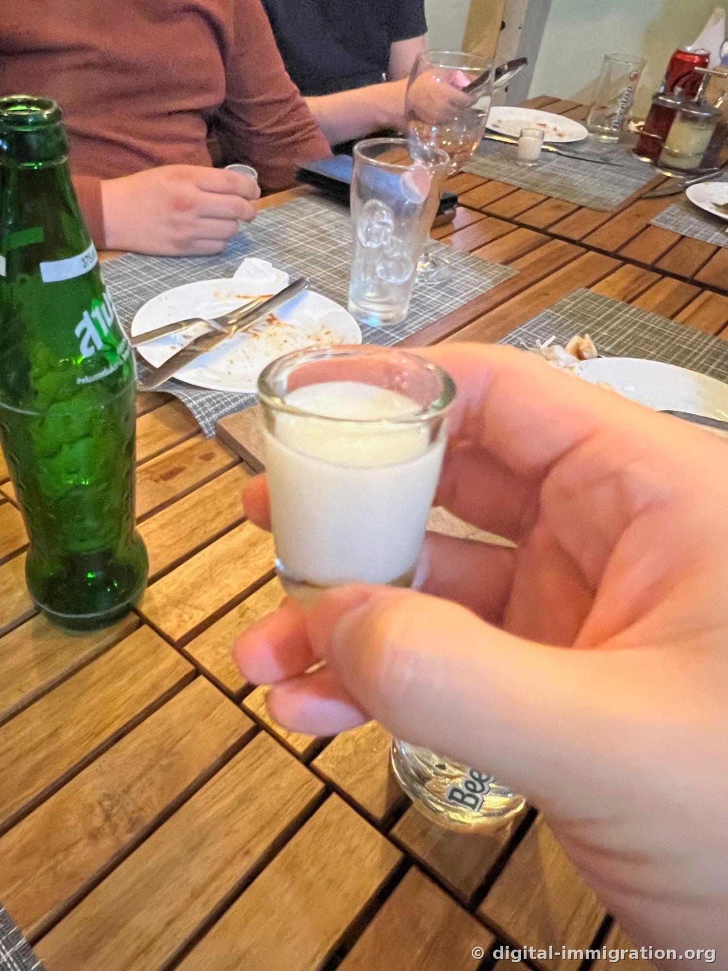 A small glass shot glass held in hand, filled with a hazy white liqueur. In the background, a green bottle of Beerlao and finished plates sit on a wooden table