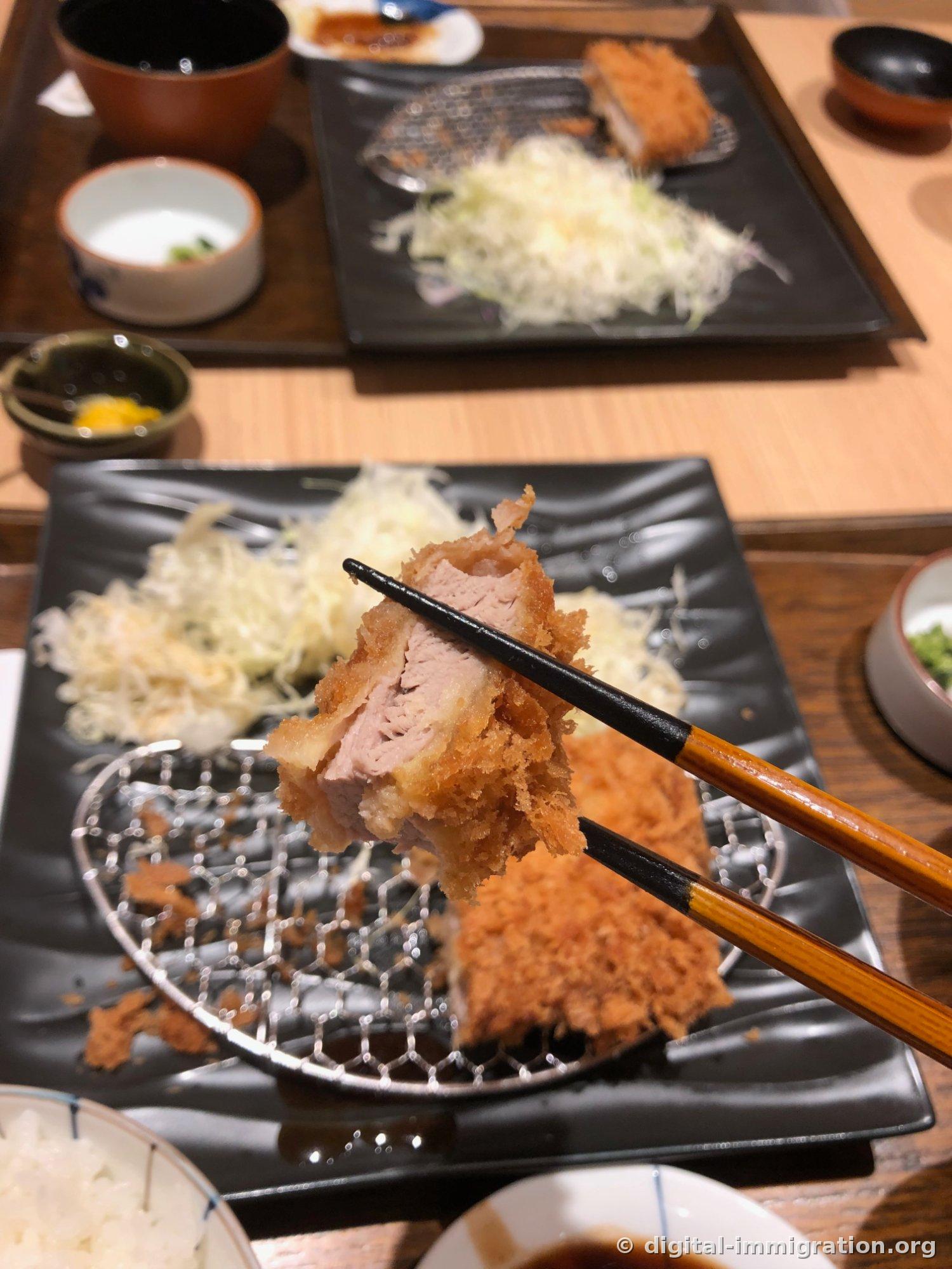 Section transversale d'une tranche de tonkatsu tenue avec des baguettes. Viande rose pâle avec couche de gras sur le bord et pâte à frire épaisse