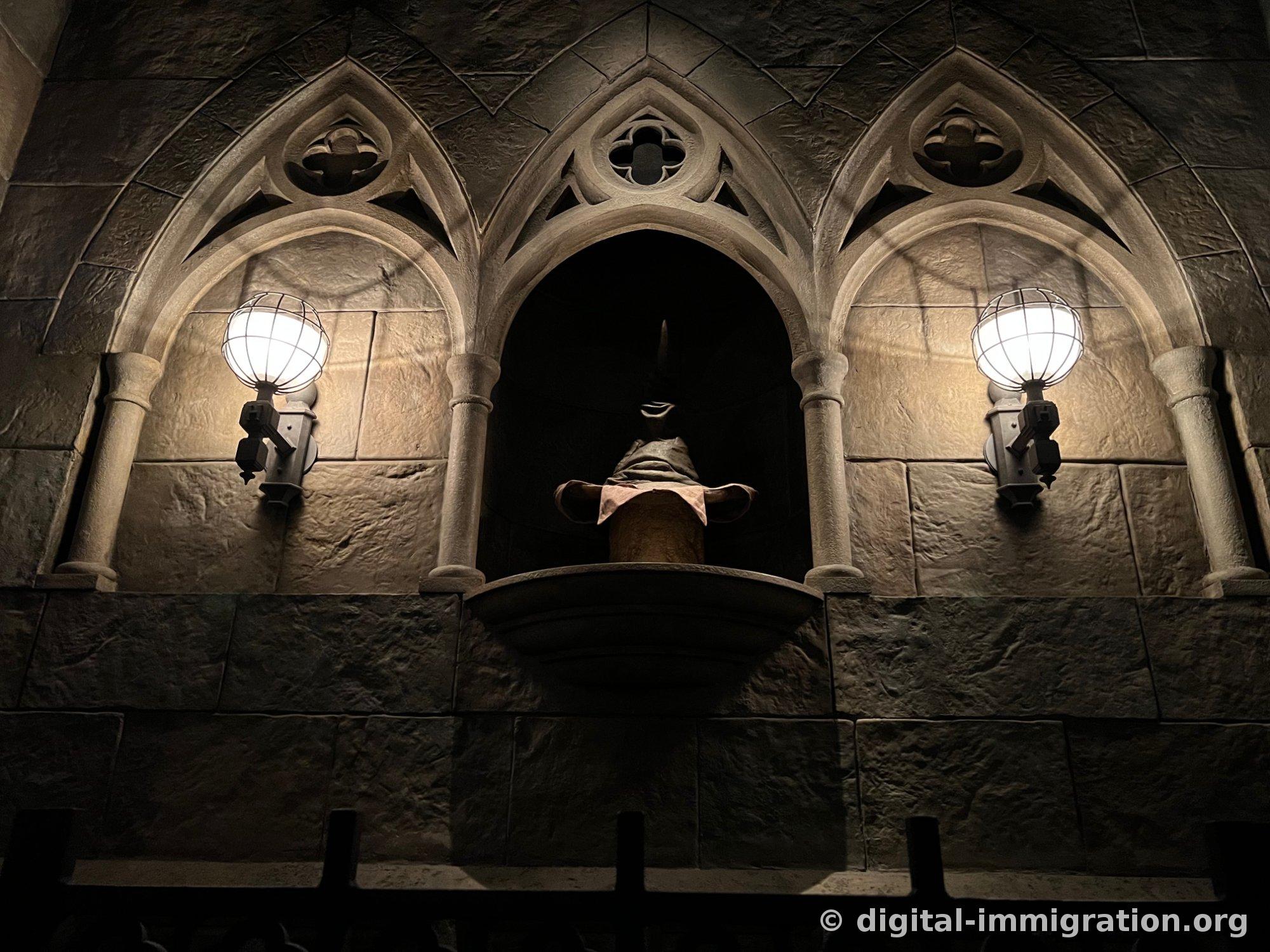 Dark stone corridor inside Hogwarts Castle, three Gothic arches in a row with a sculptured niche in the center, wire-mesh lamps on either side casting yellow light
