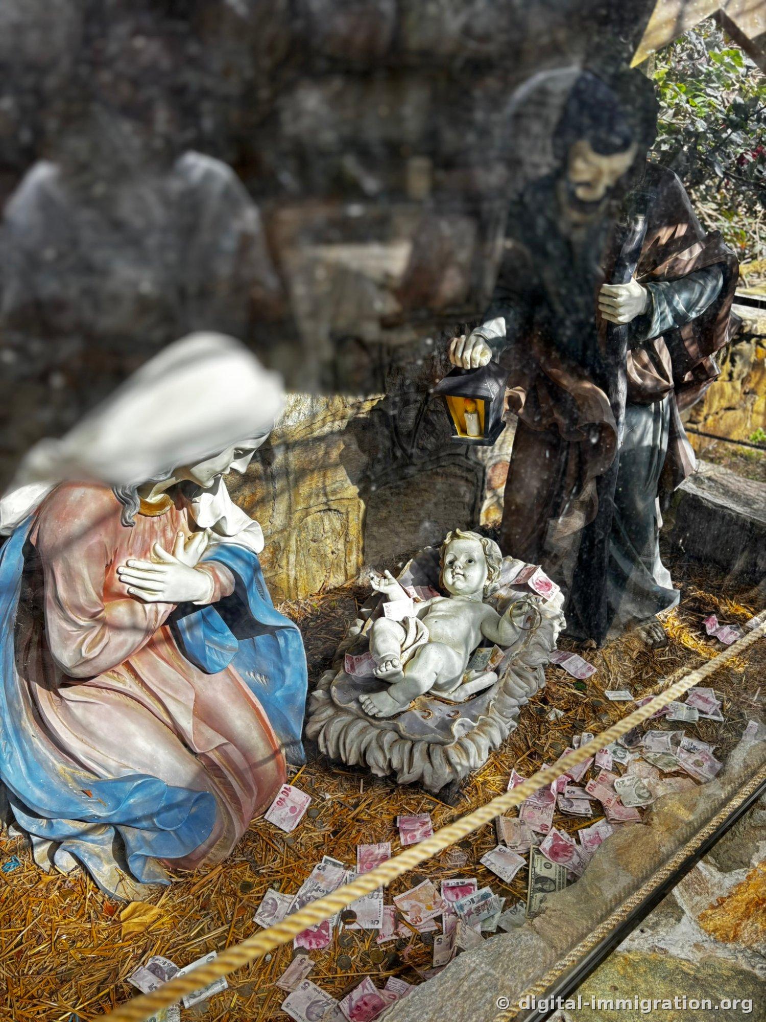 A life-size nativity scene visible through glass: Mary kneeling, baby Jesus on a shell-shaped cradle, Joseph with a lamp, and lira bills and coins scattered over straw on the floor