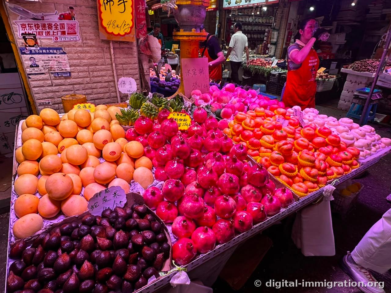Under pink lighting: yellow pomelo, finger-like Buddha's hand, piles of red pomegranates, mesh-netted flat peaches, and Indonesian salak, with a vendor in red apron drinking water