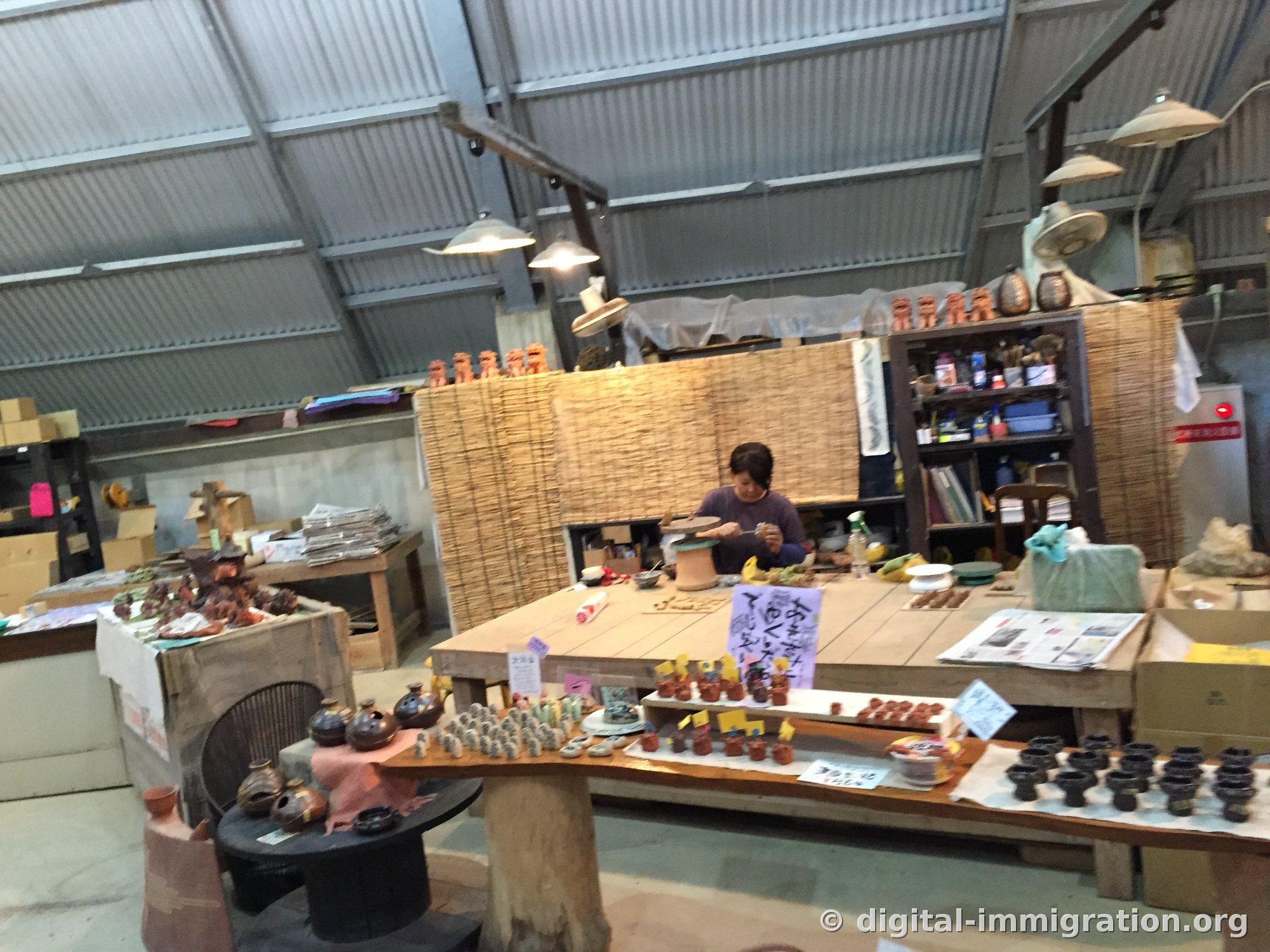 À l'intérieur de l'atelier de céramique sous un toit de tôle : un artisan assis devant un tour de potier travaillant sur une pièce, des lions Shisa rouges alignés sur des étagères, et des poteries en vente sur l'étal