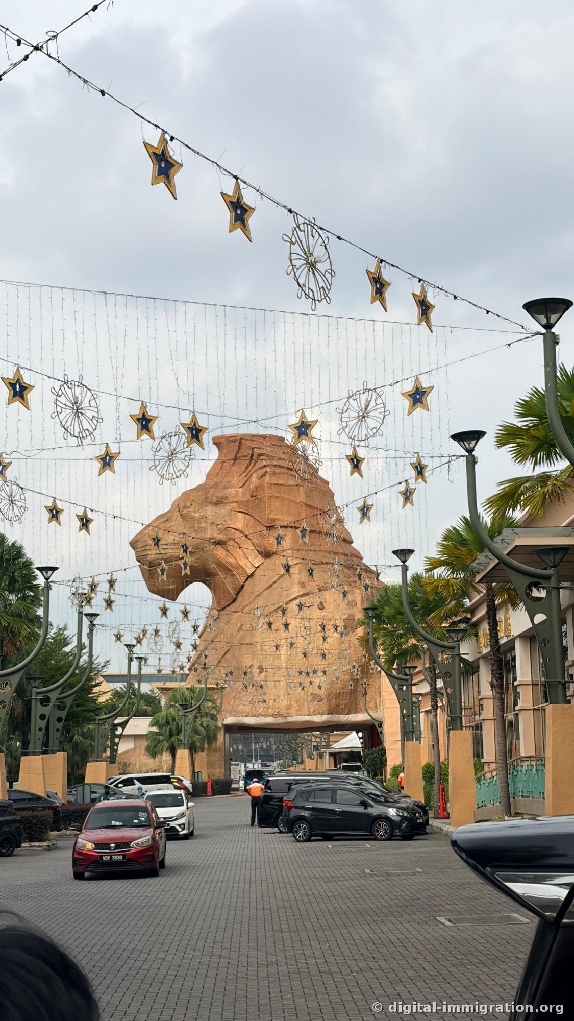 Sunway Pyramid 正门前巨大的砂岩色狮子头雕像和马路上悬挂的星形装饰，下方通过的小汽车