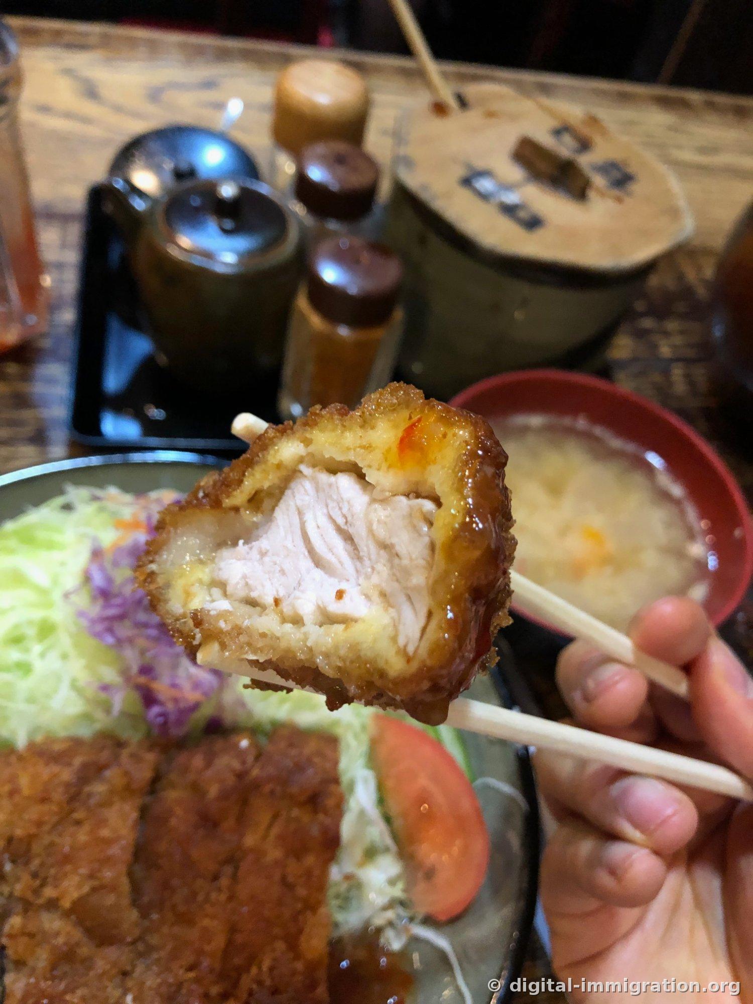 Cross-section of a fried cutlet piece held with chopsticks, with sauce on the white meat, bottles of condiments and a wooden-lidded rice container and miso soup bowl visible in the background