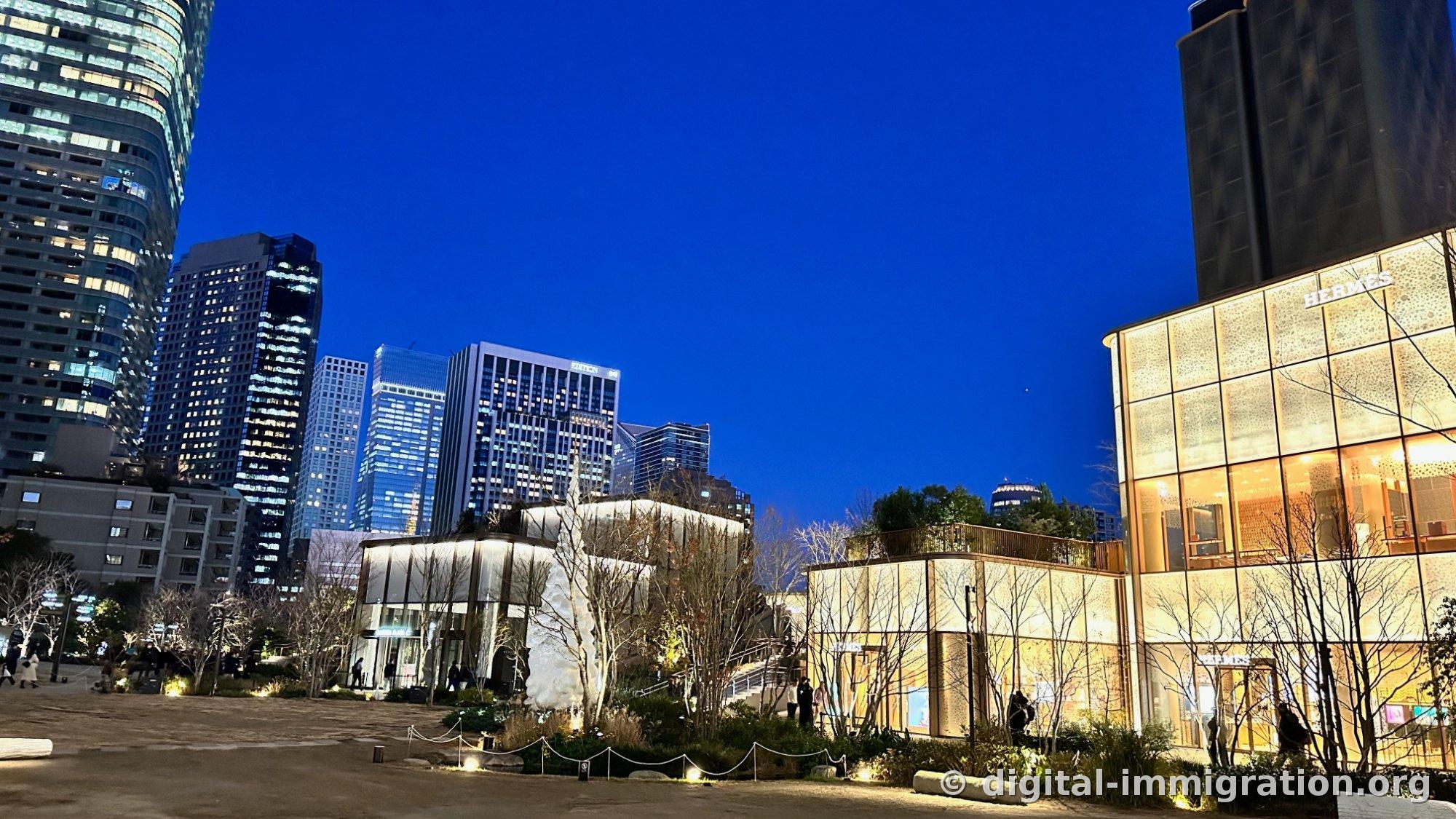 Panorama of Azabudai Hills plaza at night, with illuminated high-rise buildings on the left, white conical sculpture in the center, and Hermès store on the right