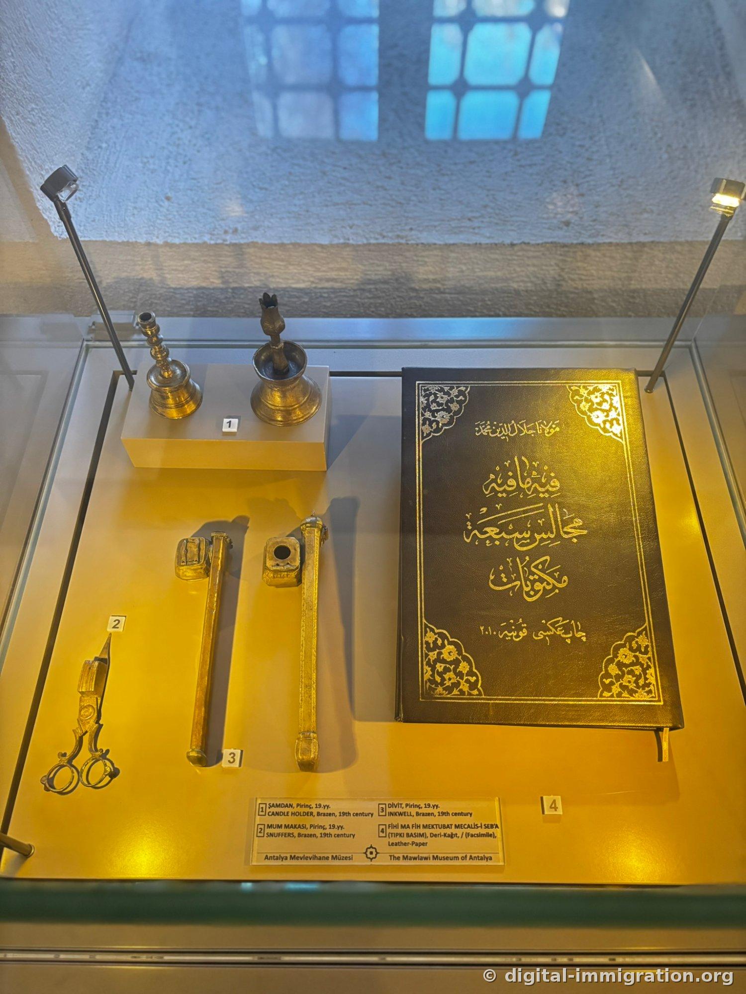 Sous l'éclairage jaune dans une vitrine en verre, chandelier en laiton du 19e siècle, ciseaux pour mèche, encrier portatif et livre à couverture noire en cuir avec caractères arabes dorés